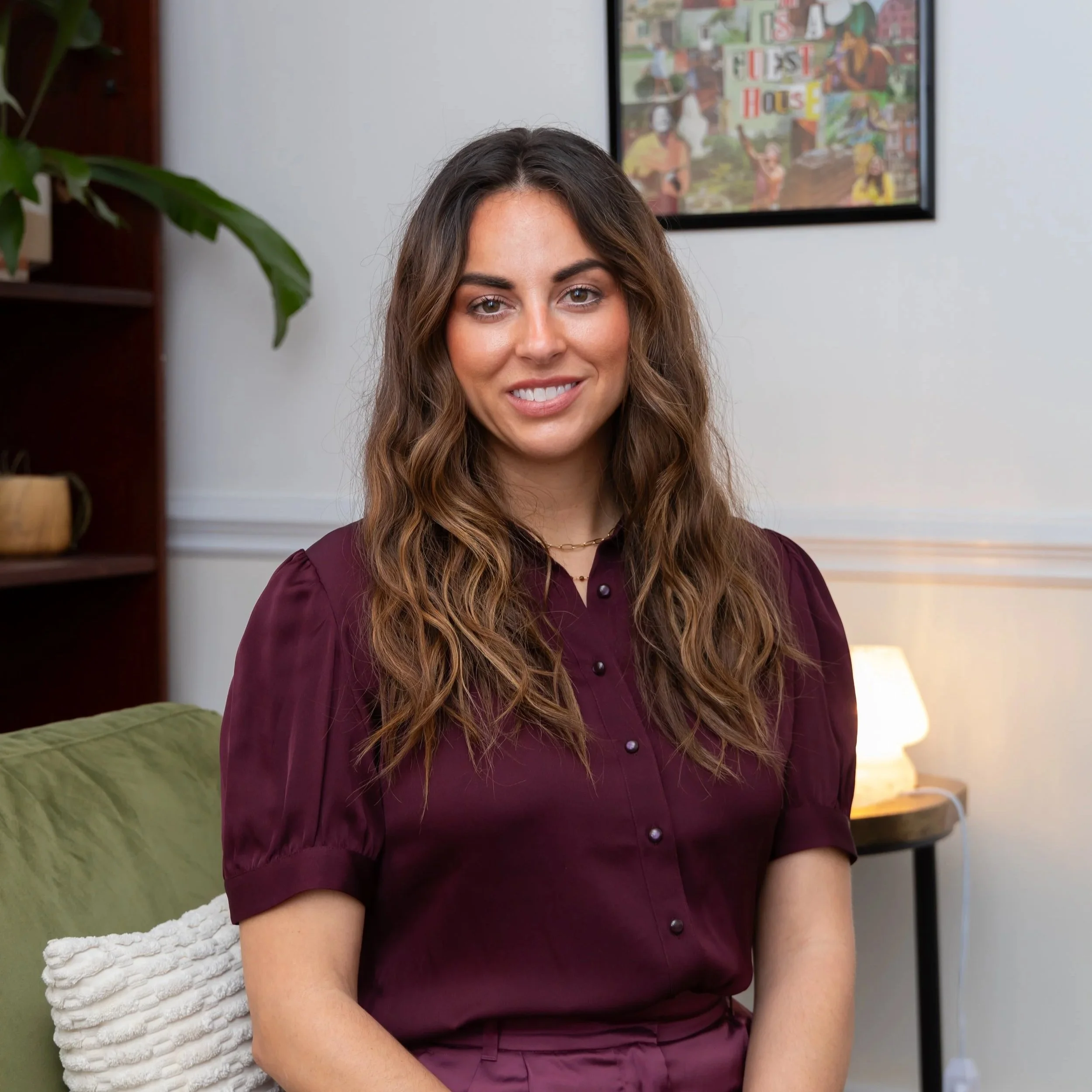 A young woman with long, wavy brown hair and light skin, smiling, sitting on a green couch with a white textured pillow. She is wearing a burgundy satin blouse and matching pants, with jewelry including a ring, bracelet, and necklace. Behind her, a framed collage poster hangs on a white wall, and a lamp provides warm lighting in the background.