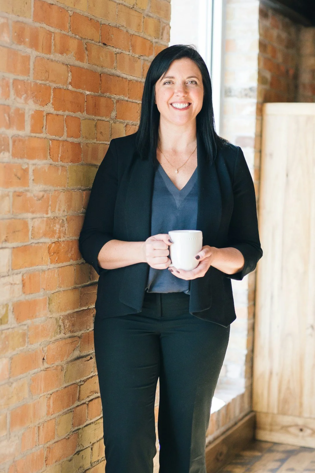 Professional therapist at Ethredge Counseling Group, with black hair and a black blazer, holding a mug against a brick wall, creating a welcoming environment for Charleston clients.