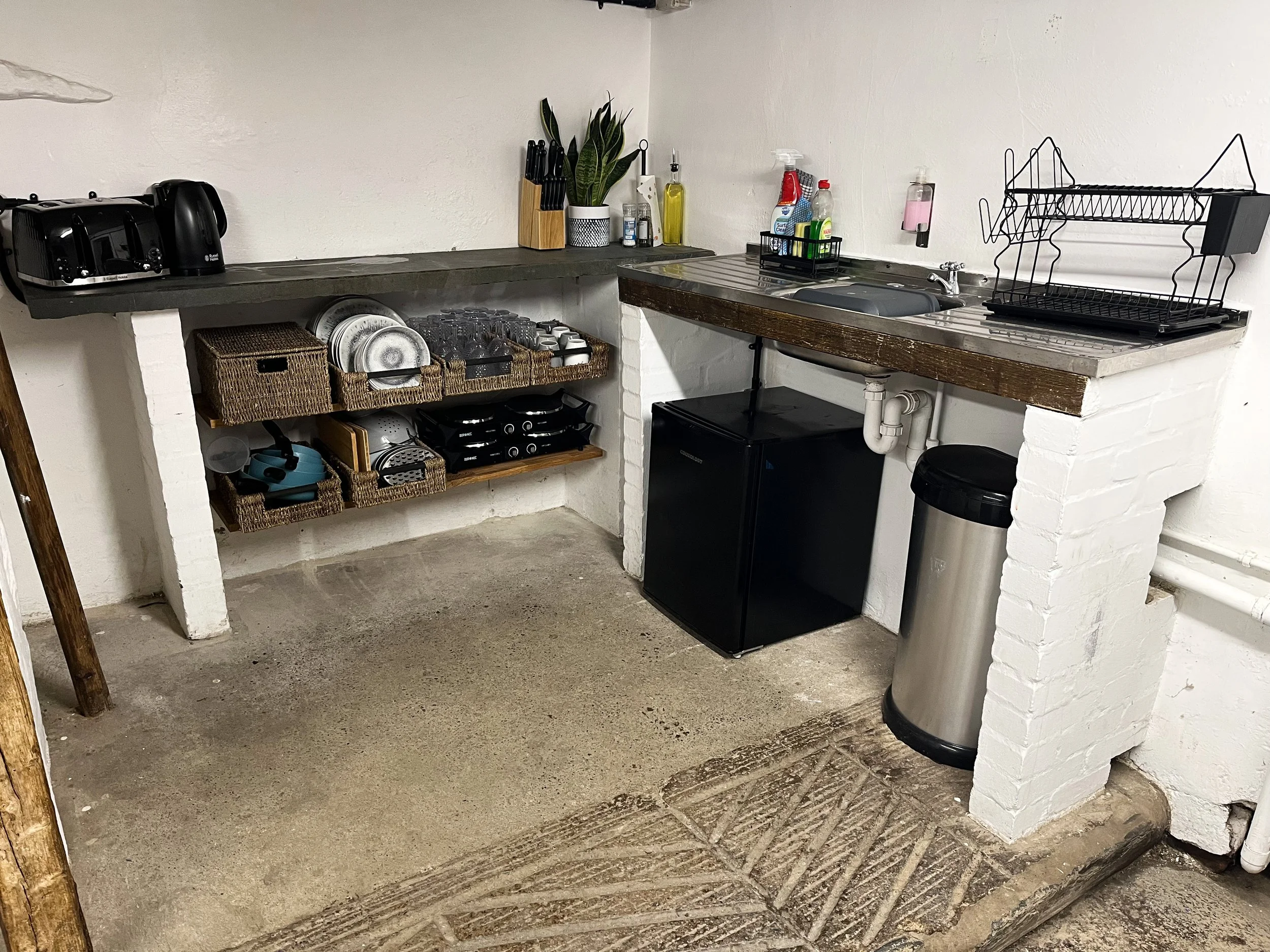 A small kitchen area with a black mini refrigerator, a sink, a metal dish rack, and various kitchen utensils and cleaning supplies on the countertops. There are baskets with dishes, cups, and kitchen tools underneath the counter, and a trash can next