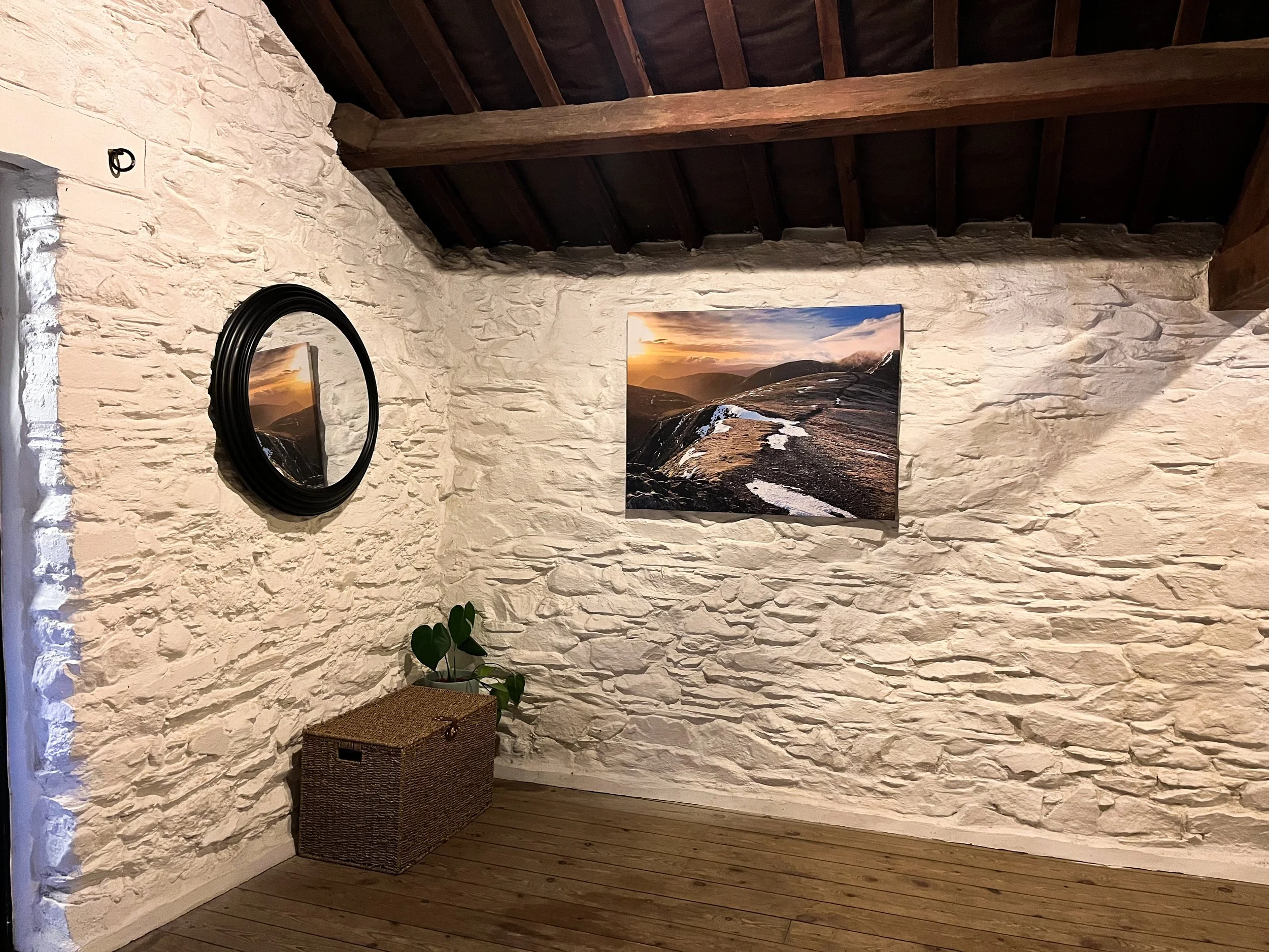 Interior of a rustic room with white stone walls, wooden ceiling beams, a framed landscape photograph, a black circular mirror, a wicker basket, and a green houseplant.