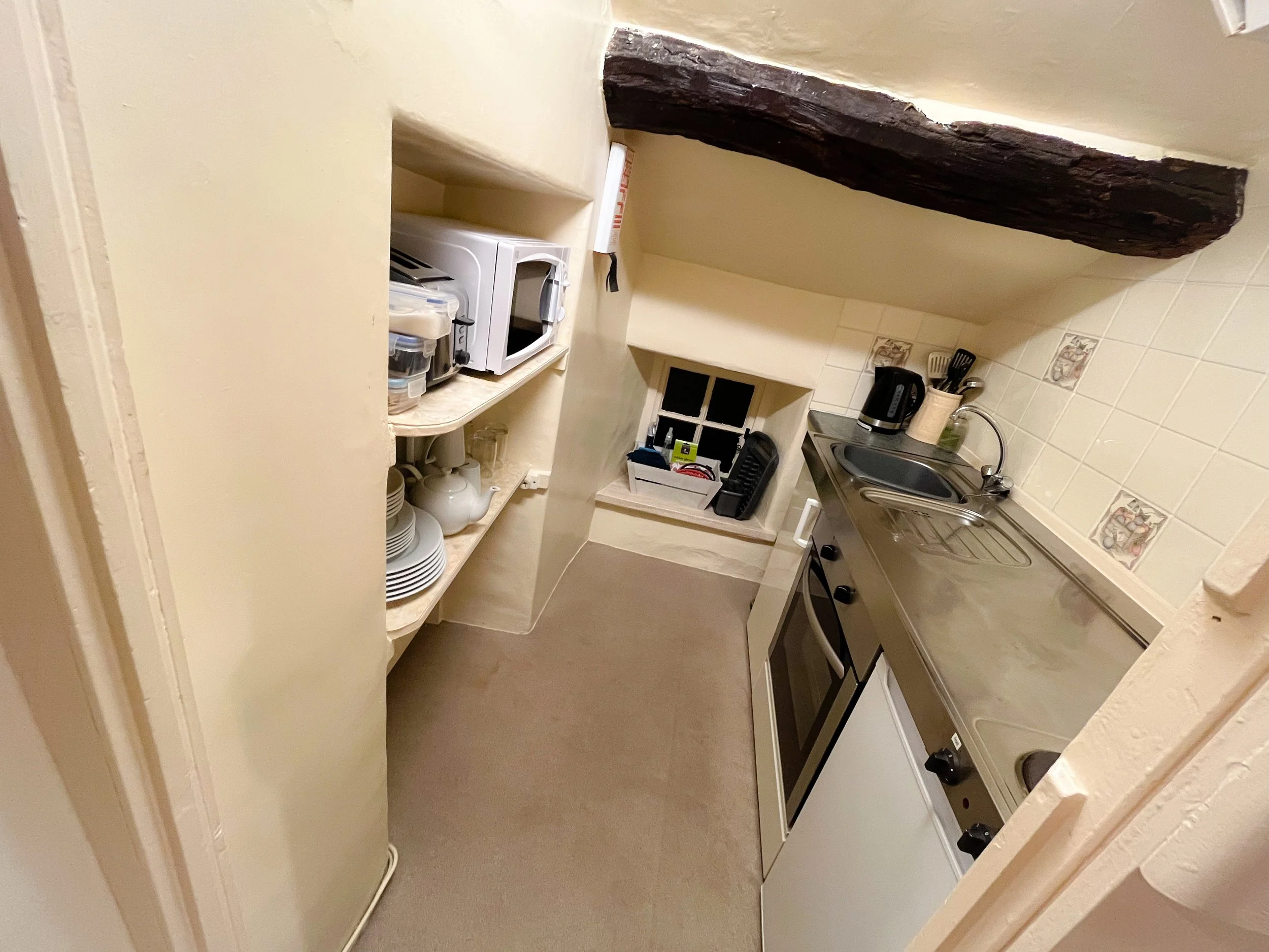 Small kitchen with beige walls and a sloped ceiling. Features a black wooden beam, a compact oven, a small sink, and a window with a ledge. Shelves with dishes, a microwave, a kettle, and cooking utensils are visible.