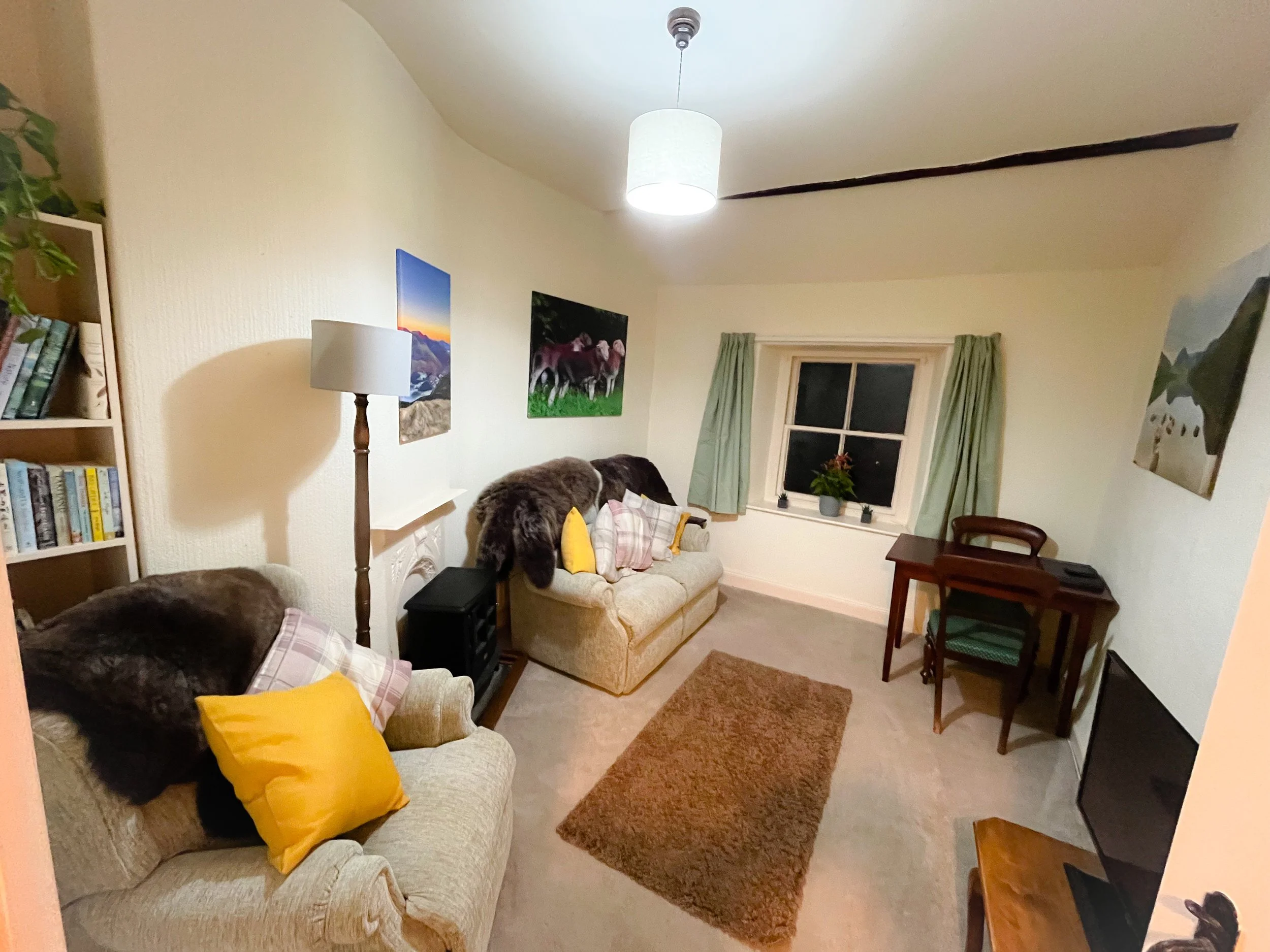 A cozy living room with beige sofas, colorful pillows, a bookshelf, a floor lamp, and framed paintings on the wall. There is a small wooden desk with a chair, a window with light green curtains, and a potted plant on the windowsill. The ceiling has a