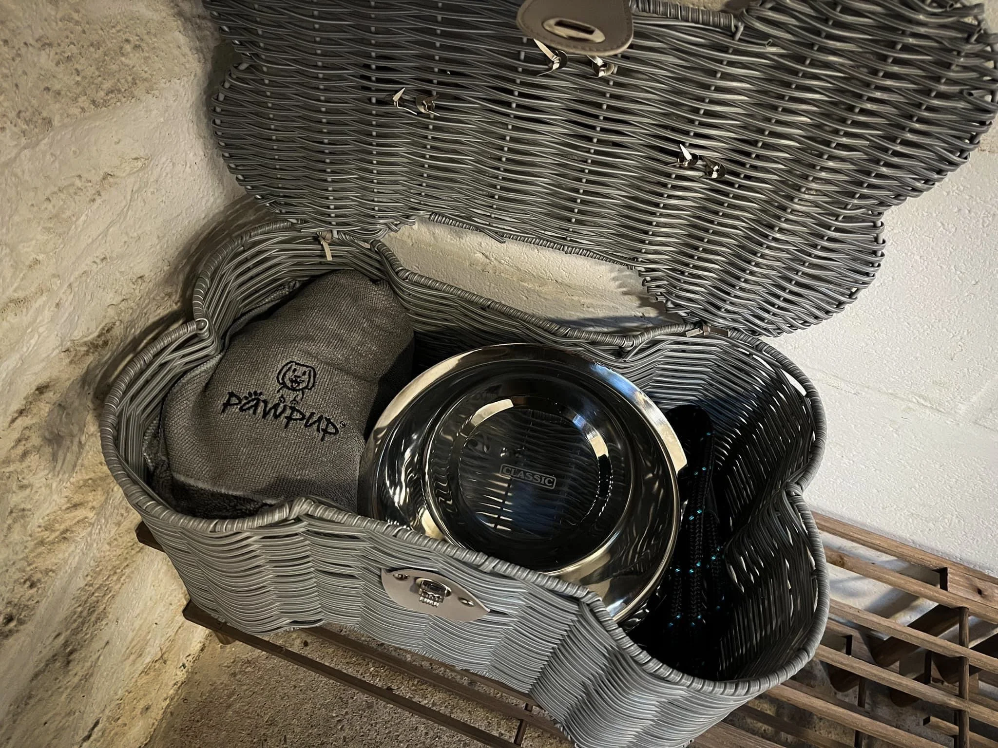 Open wicker picnic basket containing a gray drawstring bag, a chrome dog bowl, and a black leash or strap, placed on a metal rack against a stone wall and white-painted wall.