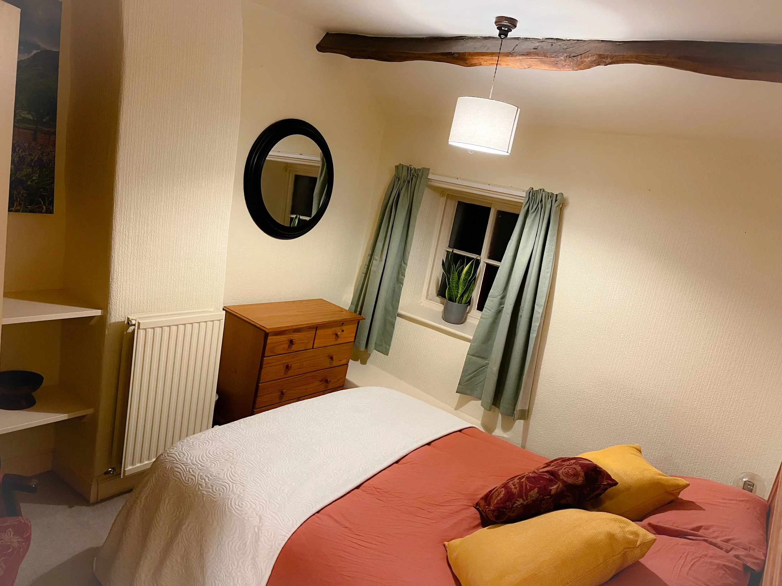 A cozy bedroom with a bed covered in a salmon-colored duvet, yellow and patterned pillows, a wooden dresser, a black oval mirror, a window with green curtains, a small potted plant on the windowsill, and a ceiling light with a fabric shade.