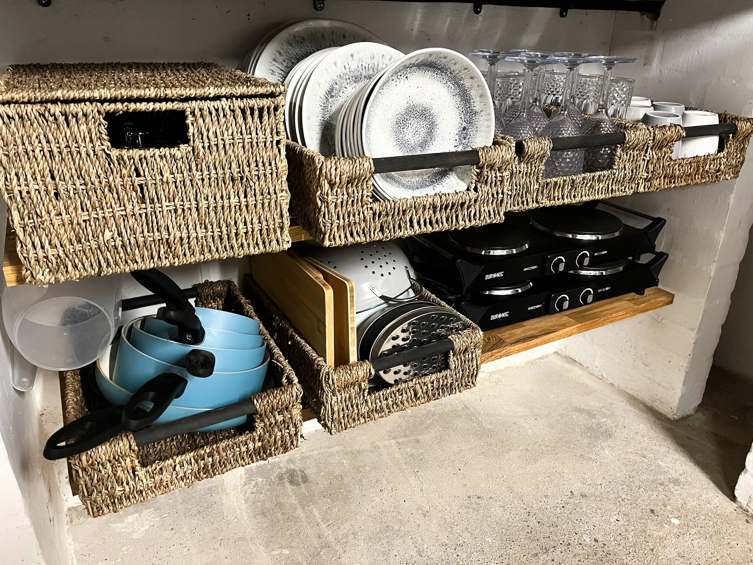 Kitchen shelving with woven baskets containing bowls, cutting boards, and a grater, plates, glasses, teacups, and various kitchenware arranged neatly.