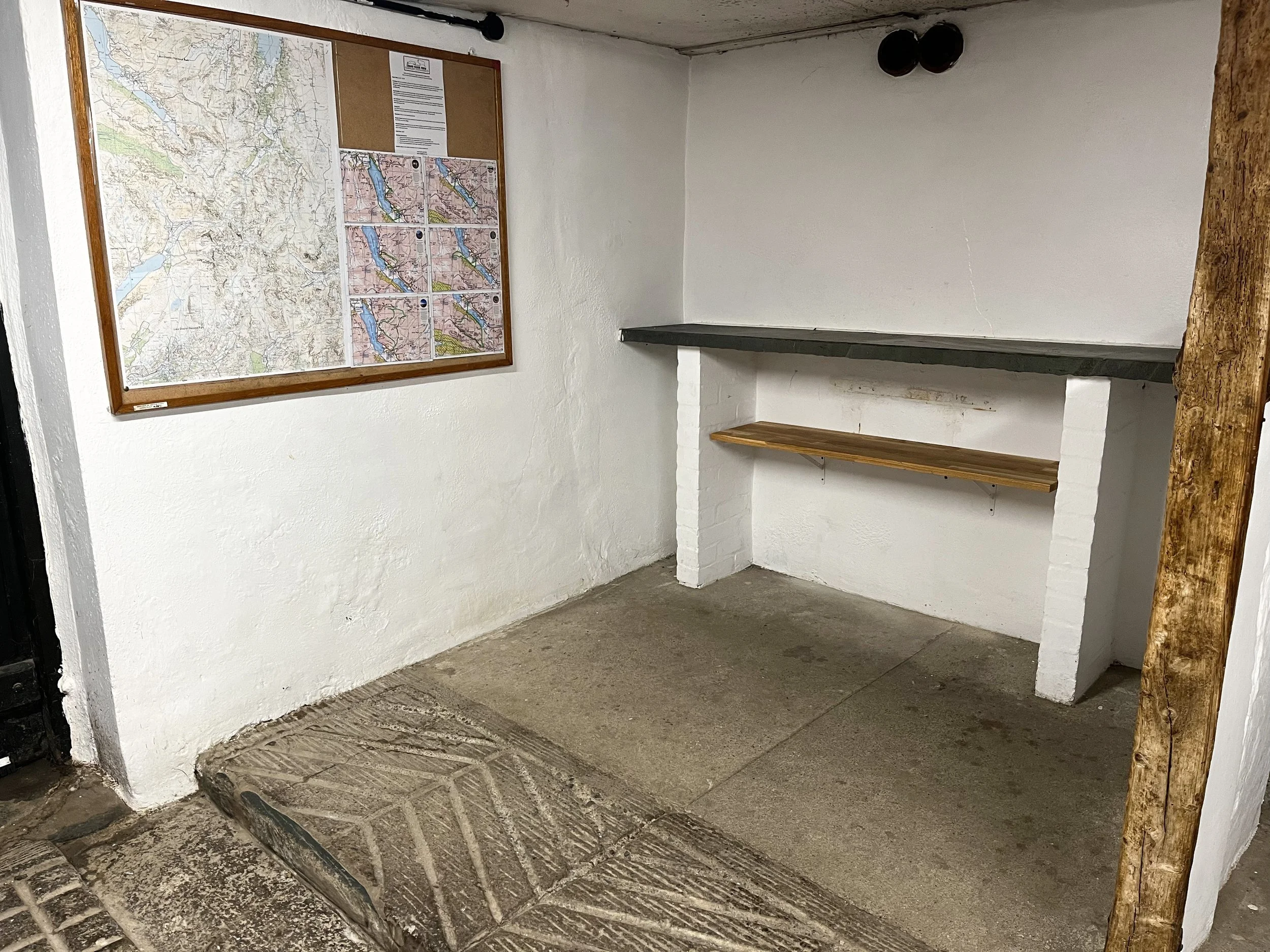 A small room with white painted brick walls, a large map on the wall, a corkboard with papers, a wooden shelf under a dark countertop, and an uneven concrete floor with a metal grate near the doorway.