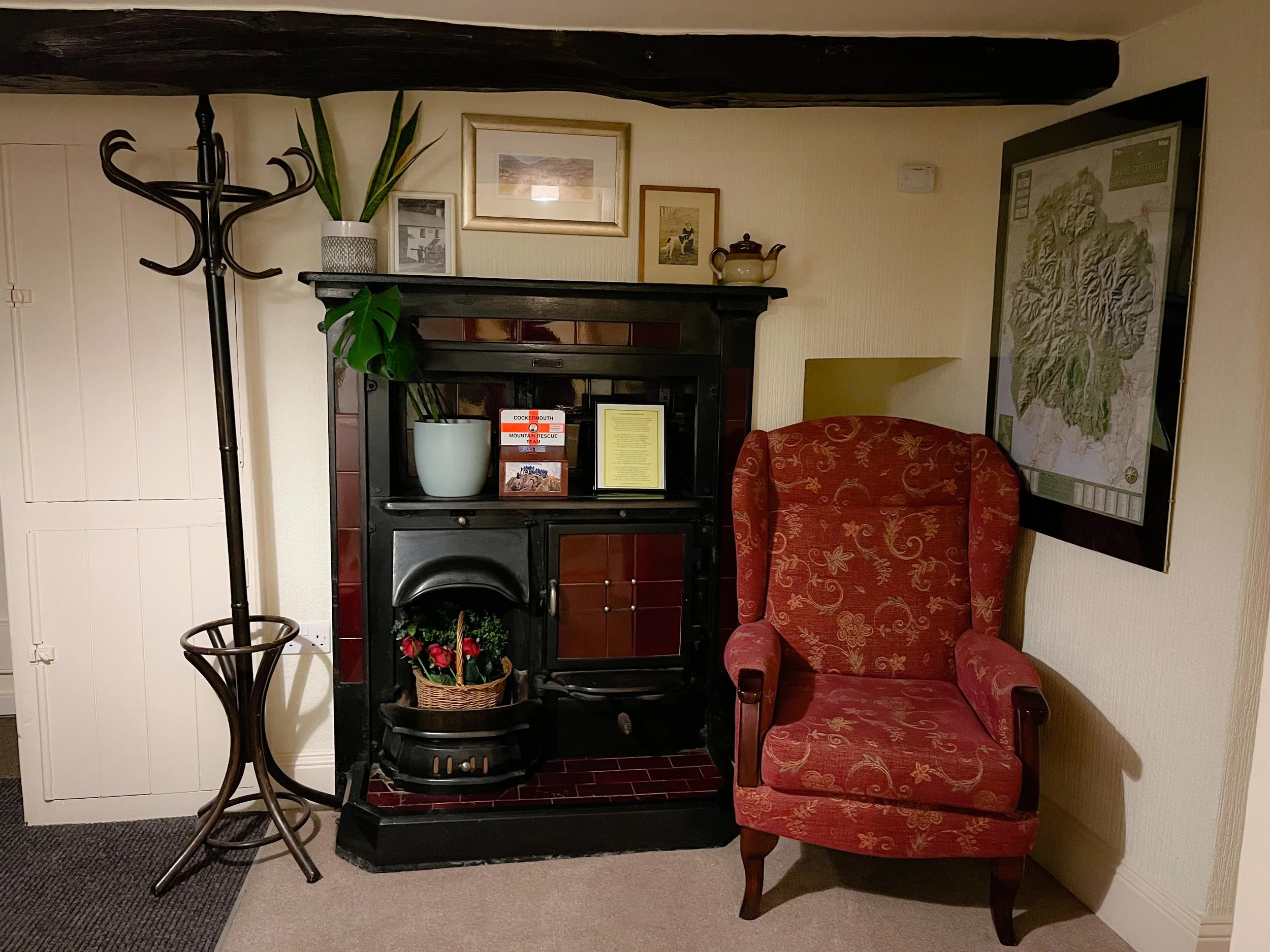 Living room corner with a red upholstered armchair, a black wood stove, a coat rack, potted plants, framed artwork, and a large map on the wall.