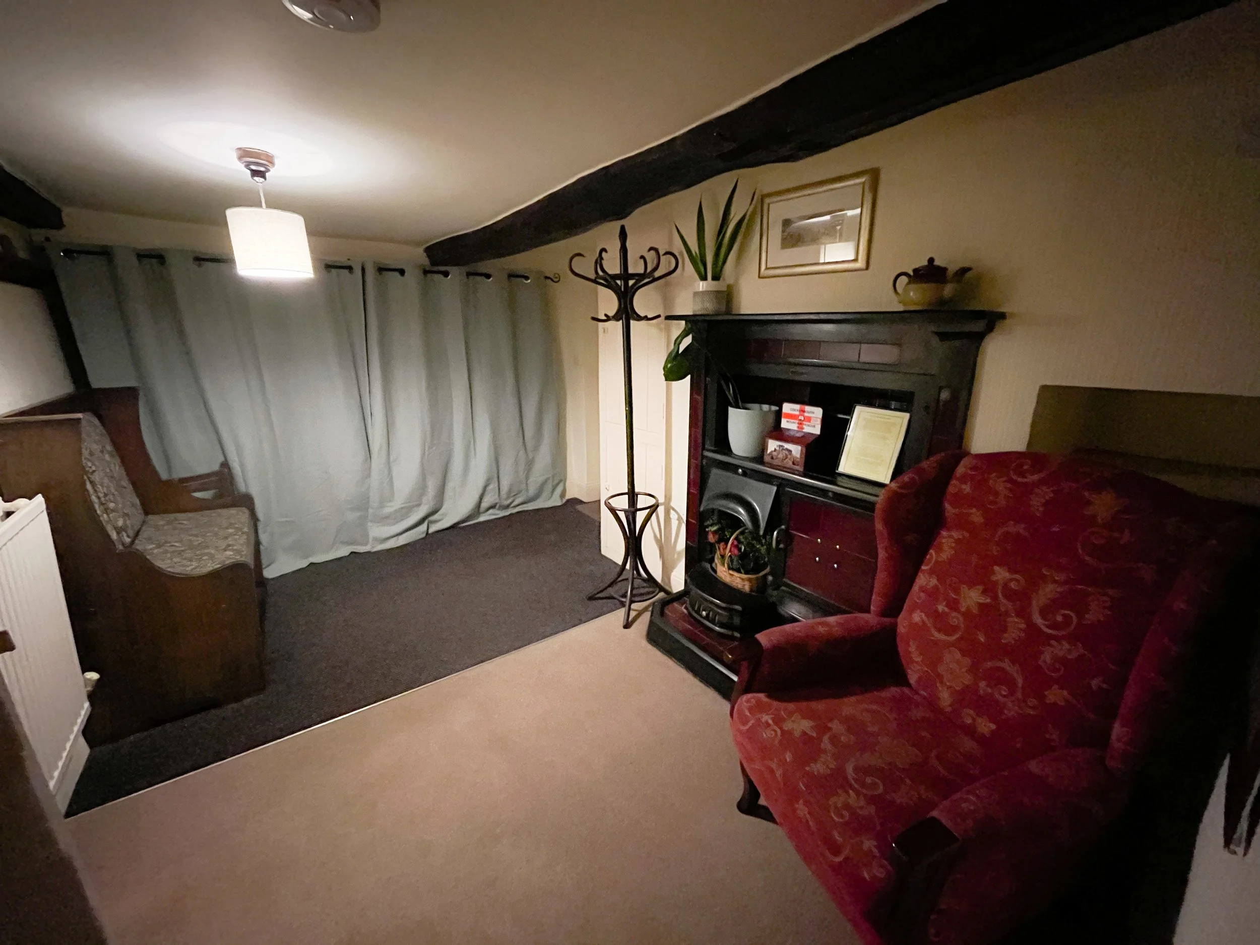 A cozy living room with a beige carpet, a red patterned armchair, a small wooden bench with floral upholstery, a black fireplace, a coat stand, a window with gray curtains, and a ceiling light fixture.
