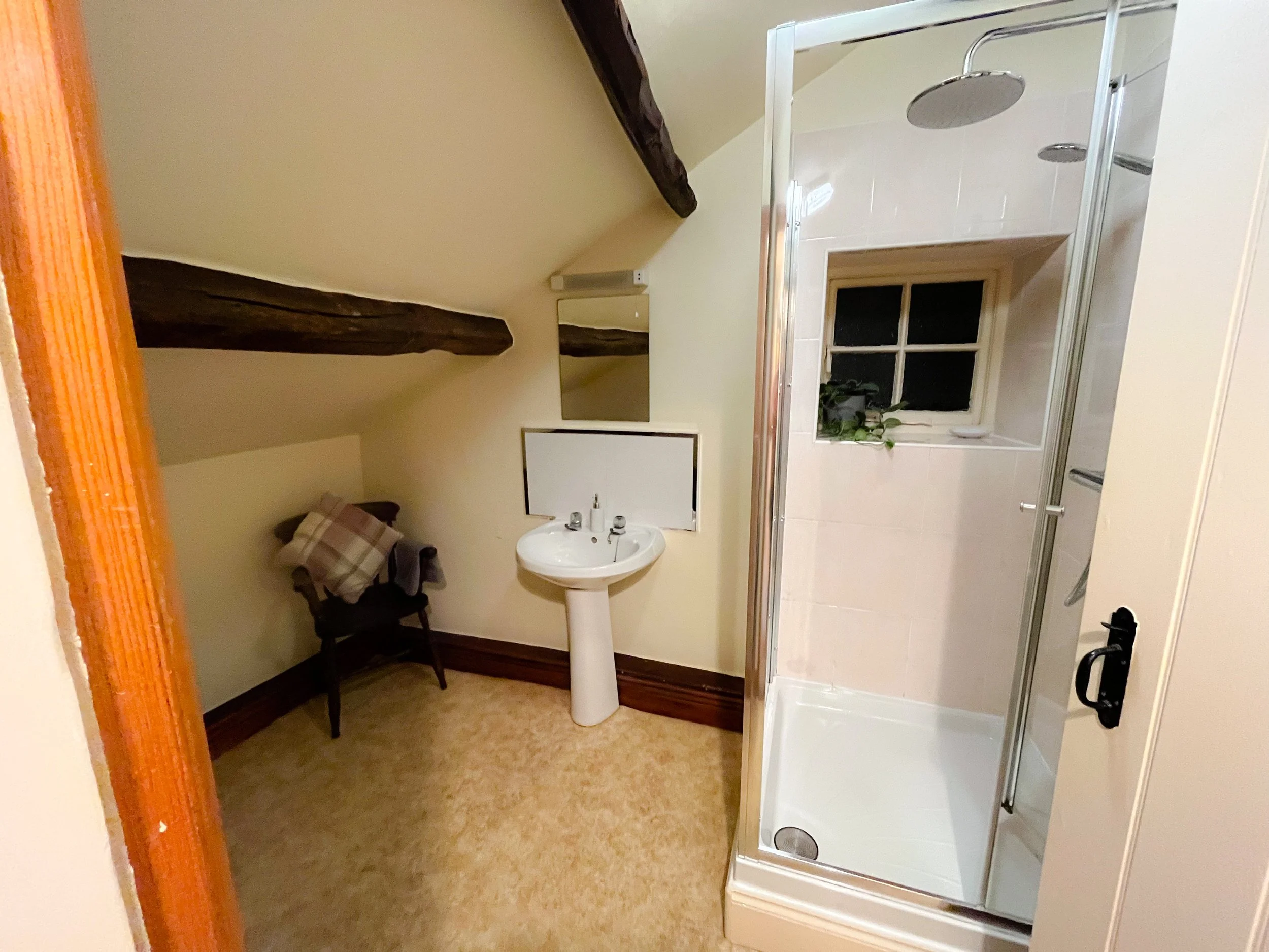 Small bathroom with a walk-in shower, a small window with a potted plant, a pedestal sink, a mirror, a wooden chair with a blanket, sloped ceiling with exposed wooden beams, beige carpet, and cream-colored walls.