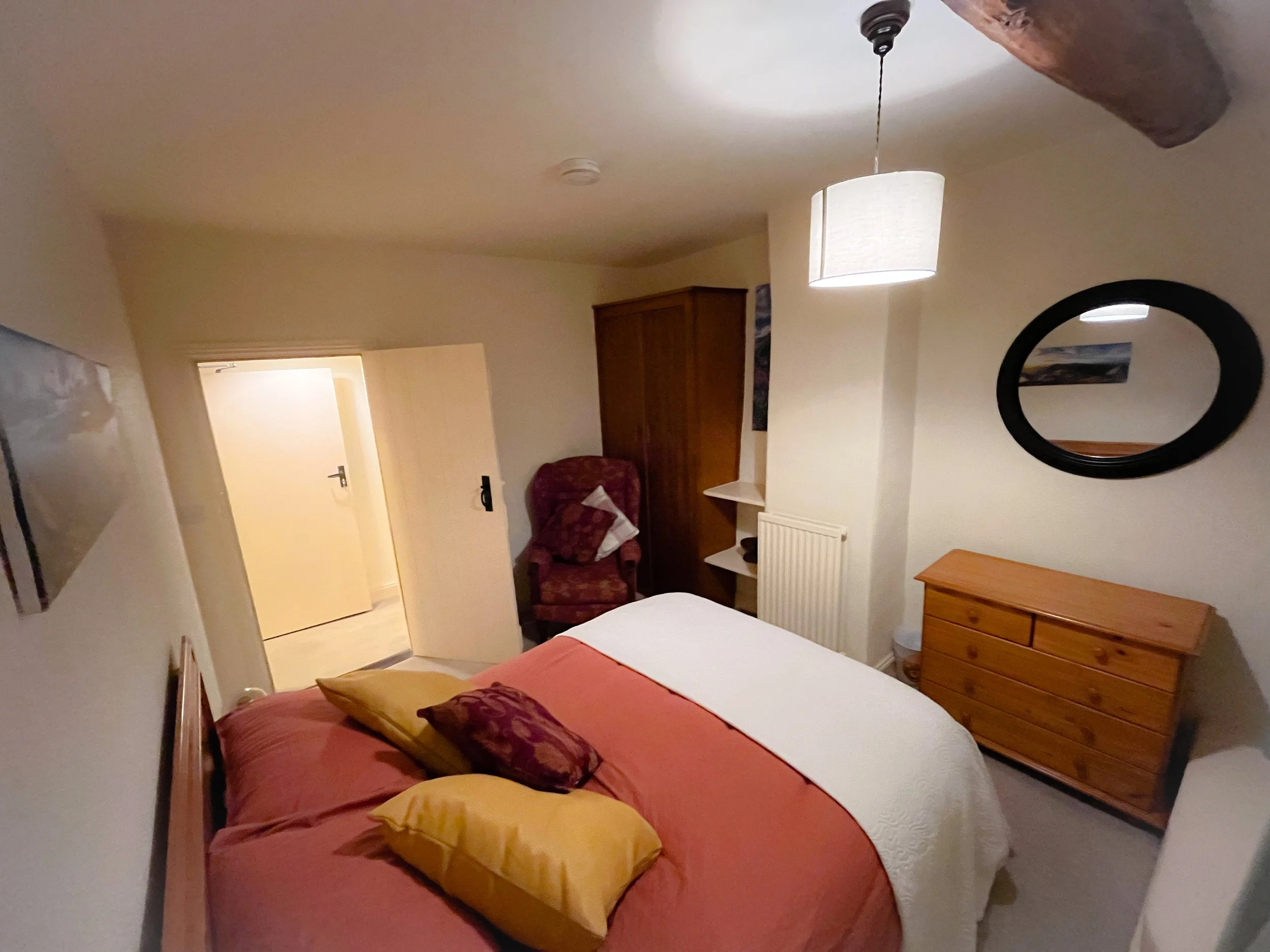 A cozy bedroom with a double bed covered in a white quilt and pink blankets, adorned with yellow and purple pillows. There is a wooden dresser on the right side, a mirror on the wall above it, and a purple upholstered armchair with a pillow near the 