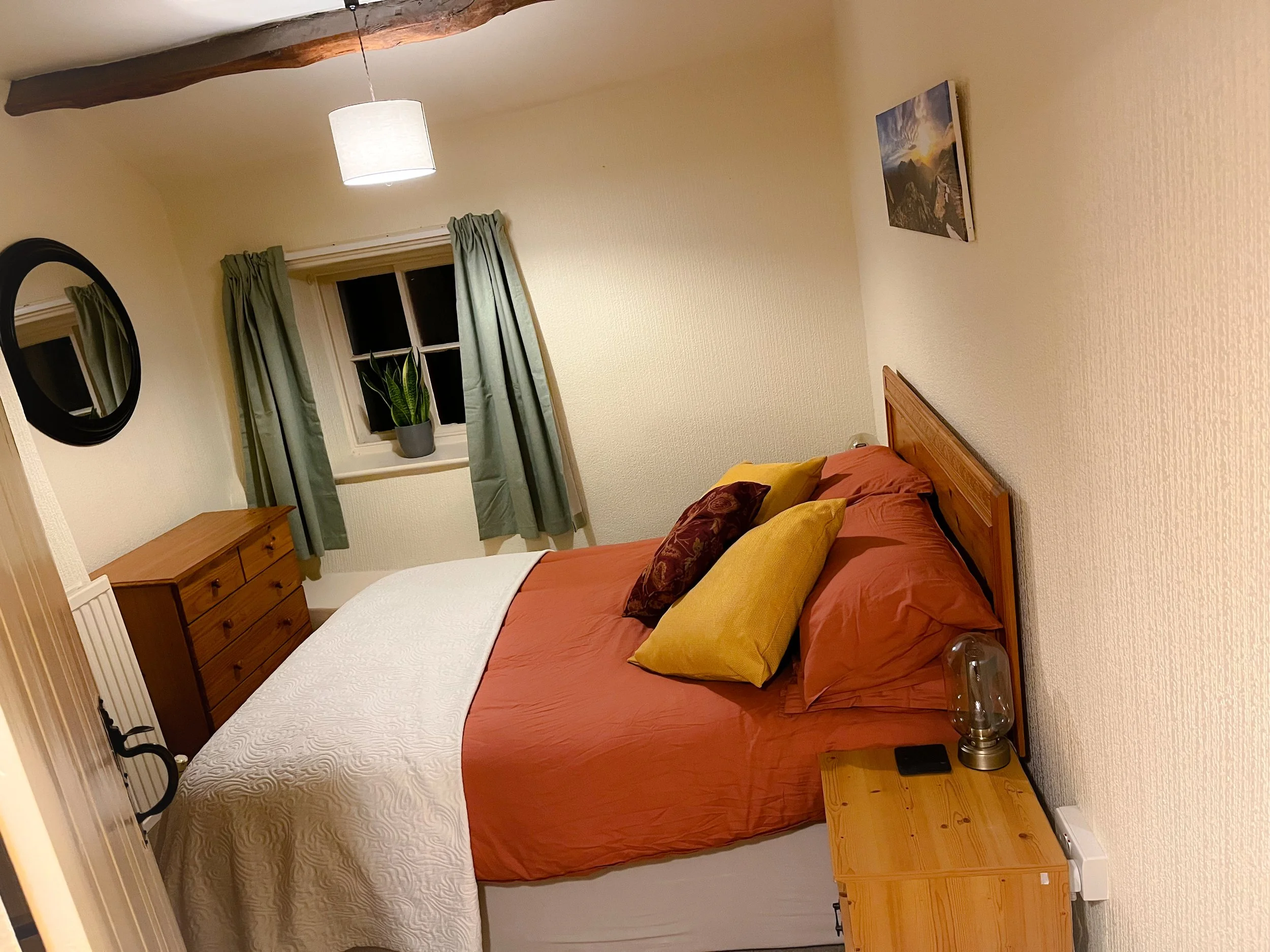 A cozy bedroom with a bed covered in earthy orange, beige, and yellow pillows, a wooden nightstand with a lamp and phone, a dresser, a mirror, a window with green curtains, a potted plant, and wall art, illuminated by a ceiling light and a rustic woo