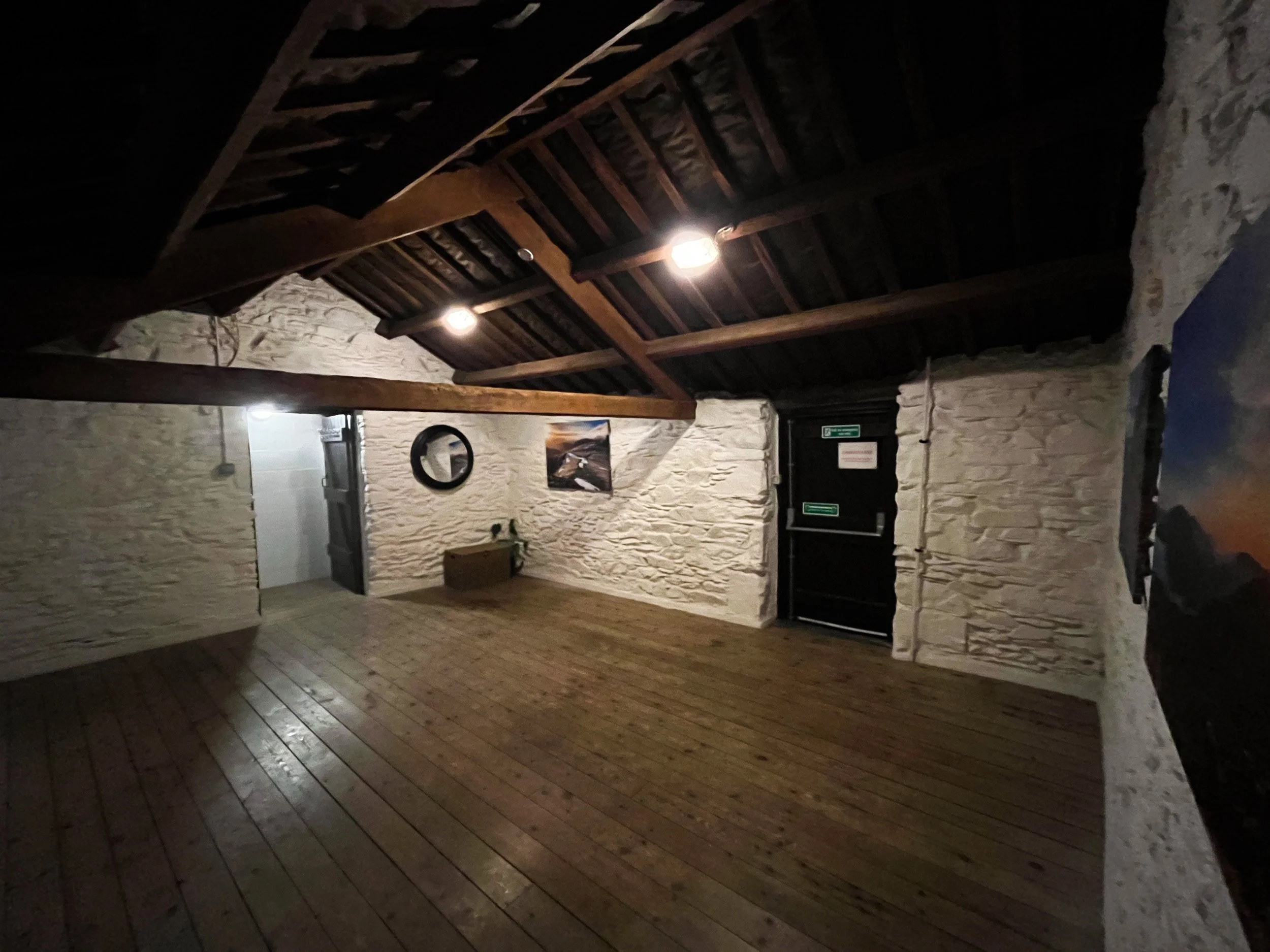Empty room with white brick walls, wooden floors, sloped wooden ceiling with exposed beams, and dim lighting. There is a small piece of furniture against the wall and some wall art.