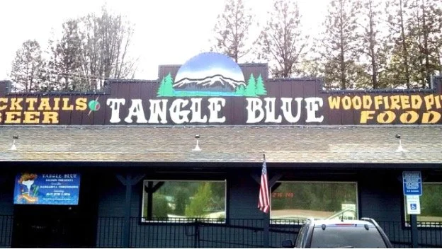 Sign for Tangle Blue bar and restaurant featuring wood-fired pizza, cocktails, beer, and food, with trees and a mountain logo in the background.