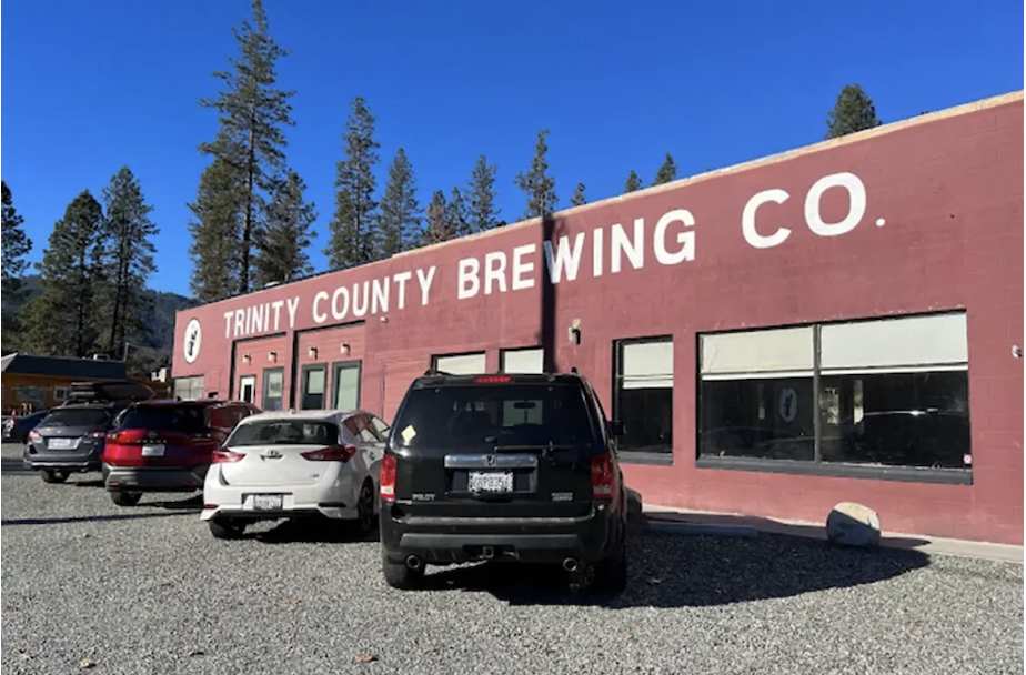 A red-brick building with large white lettering reading 'Trinity County Brewing Co.' in a parking lot filled with several parked cars, with tall trees and a clear blue sky in the background.