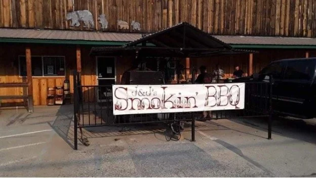 A sign on a black metal fence that reads 'J&J's Smokin' BBQ' in front of a wooden building with a green roof and a parking lot.