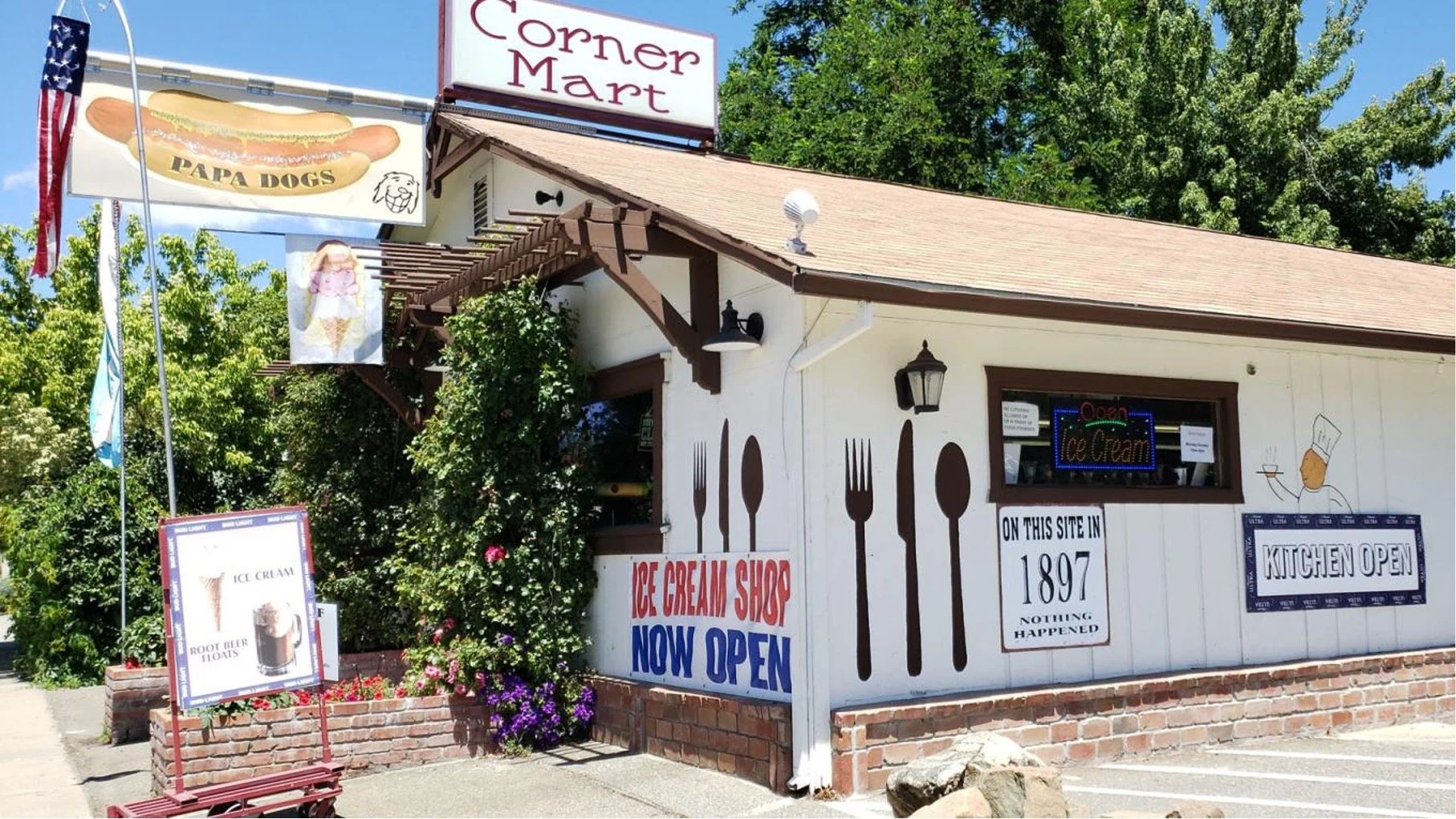An ice cream shop called Corner Mart with signs advertising hot dogs and ice cream. The shop has a white exterior with painted utensils and text announcing its opening in 1897. There are flags, flowers, and trees nearby under a blue sky.