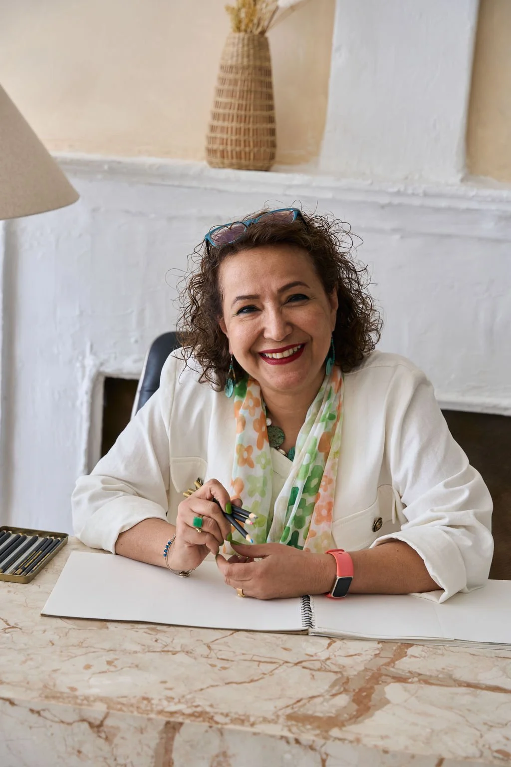 A smiling woman with curly hair, wearing a white jacket, colorful scarf, and glasses on her head, sitting at a marble desk with a notebook and colored pencils, in a cozy room with a white wall, a woven vase with flowers in the background.