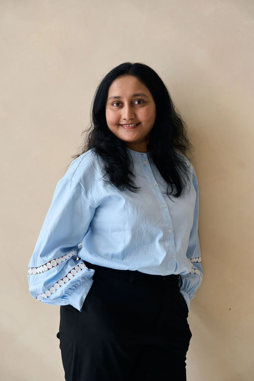 A woman with long black hair, wearing a light blue button-up shirt with decorative cutouts on the sleeves, standing against a plain beige wall and smiling.