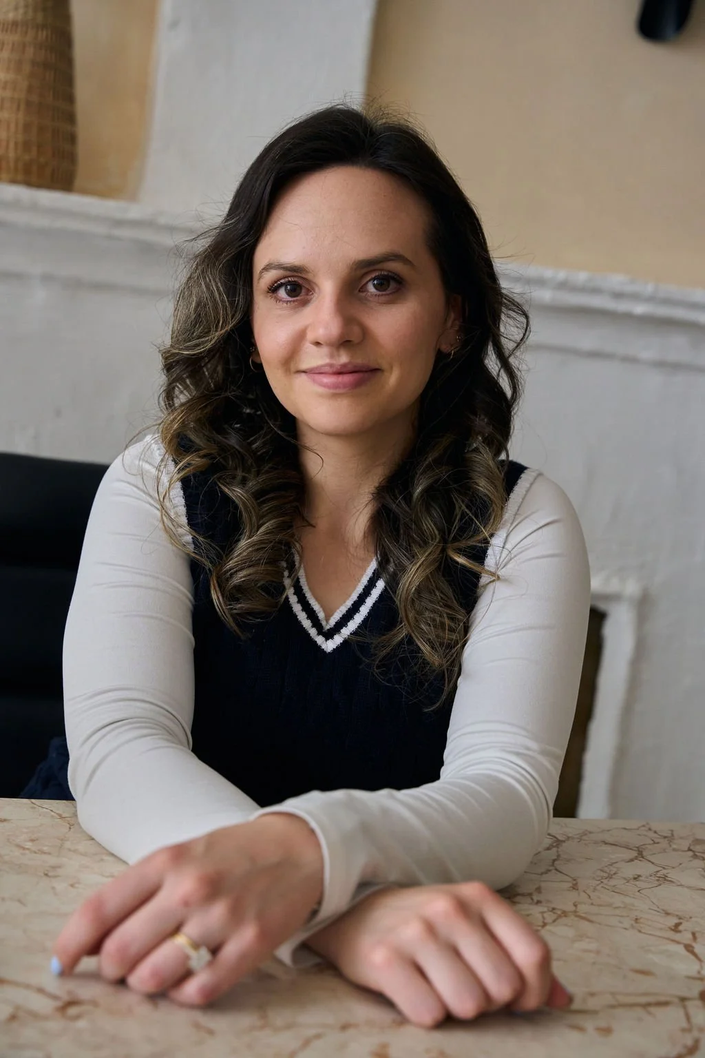 A woman with dark wavy hair sitting at a marble table in front of a white wall with a fireplace.