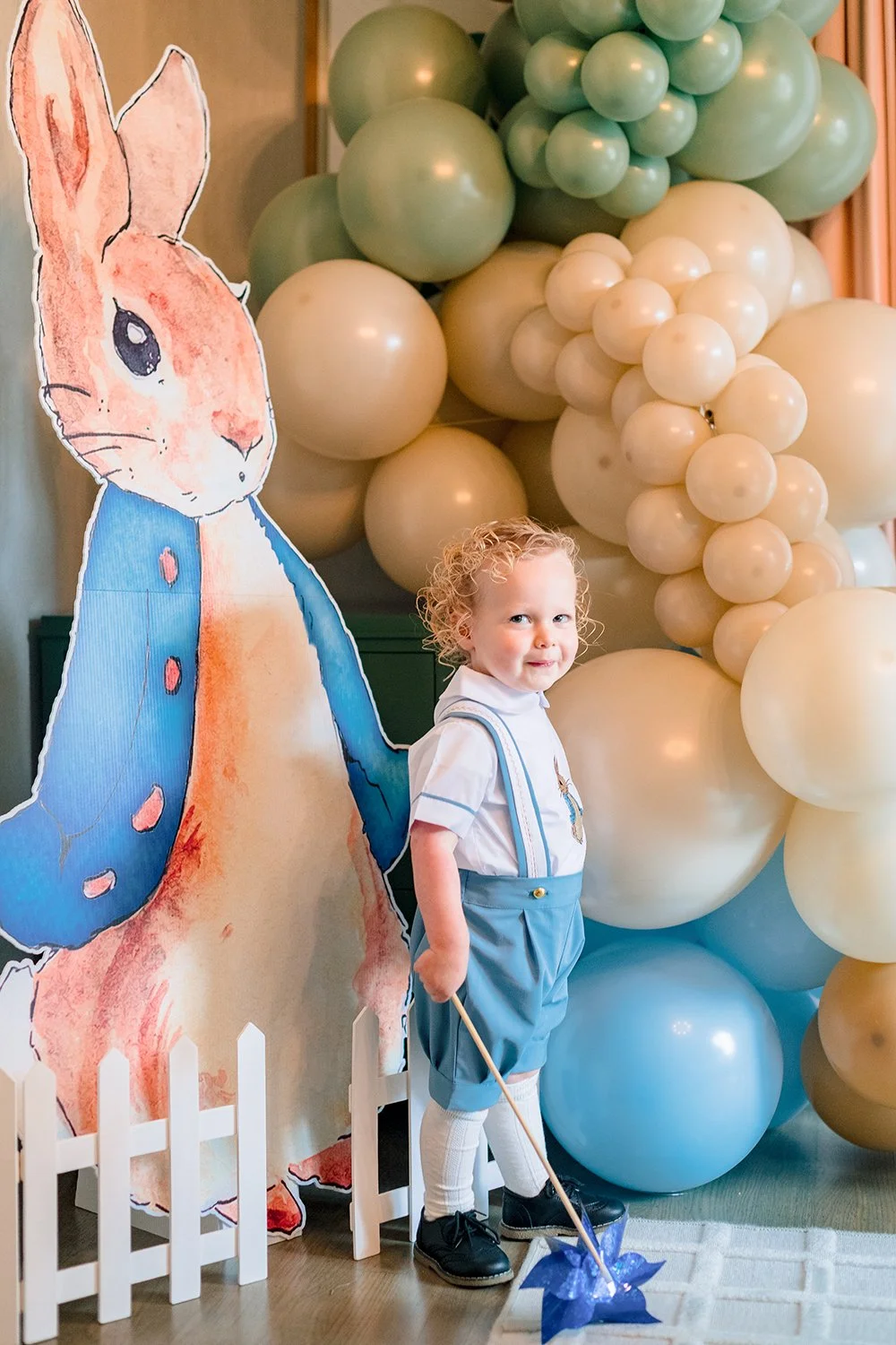 A young boy with curly blonde hair, dressed in a white shirt with blue suspenders, blue shorts, and black shoes, stands in front of a large decorative bunny cutout at a party. He is holding a small stick with a blue star-shaped decoration. Behind him