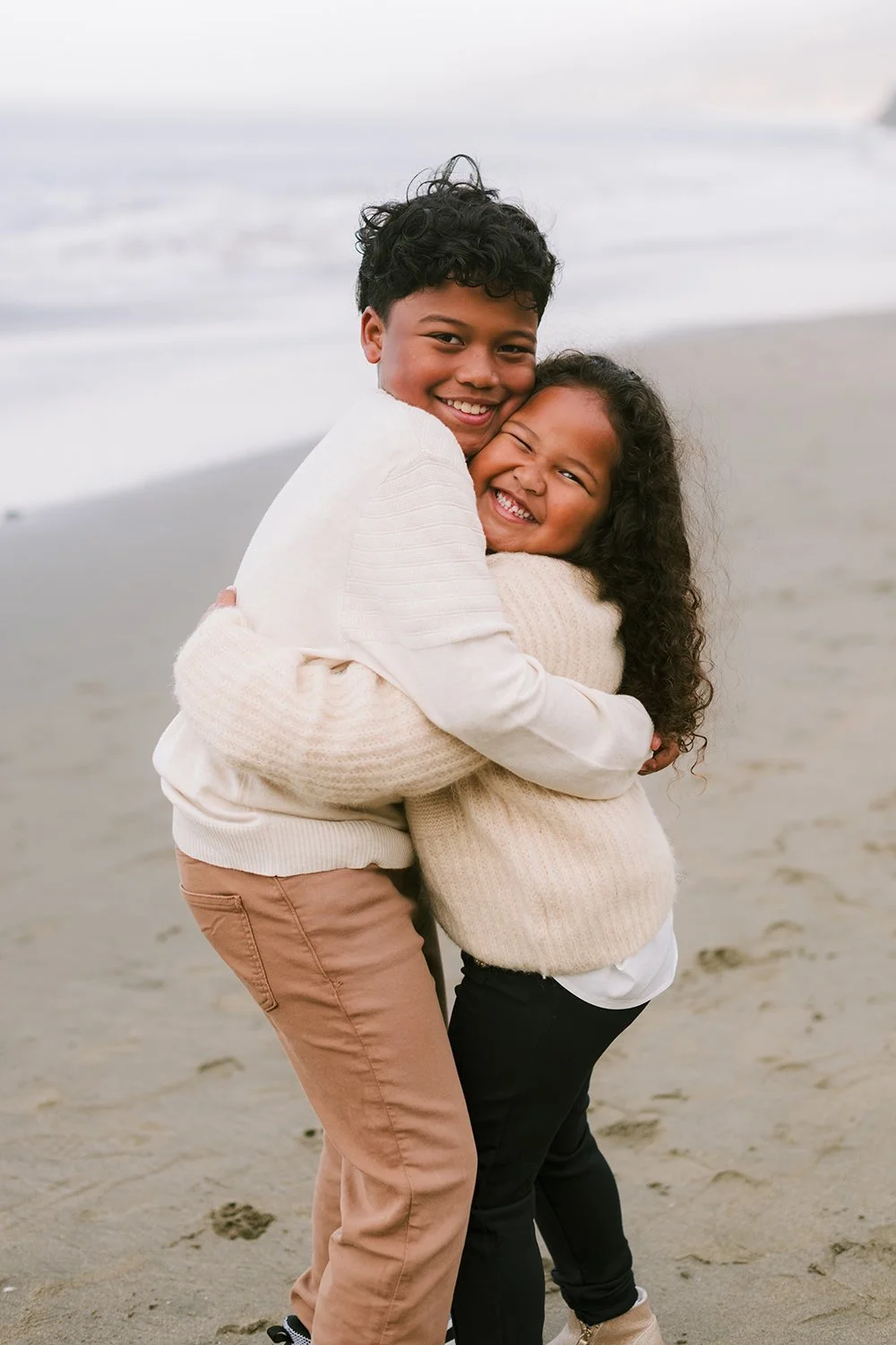 Two kids, a boy and a girl, hugging and smiling on a beach with waves in the background.