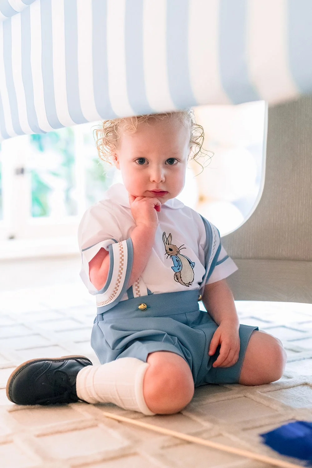 A young child with curly blonde hair, wearing a white shirt with a bunny design and blue shorts, sitting on a carpeted floor under a striped curtain, with a thoughtful expression.
