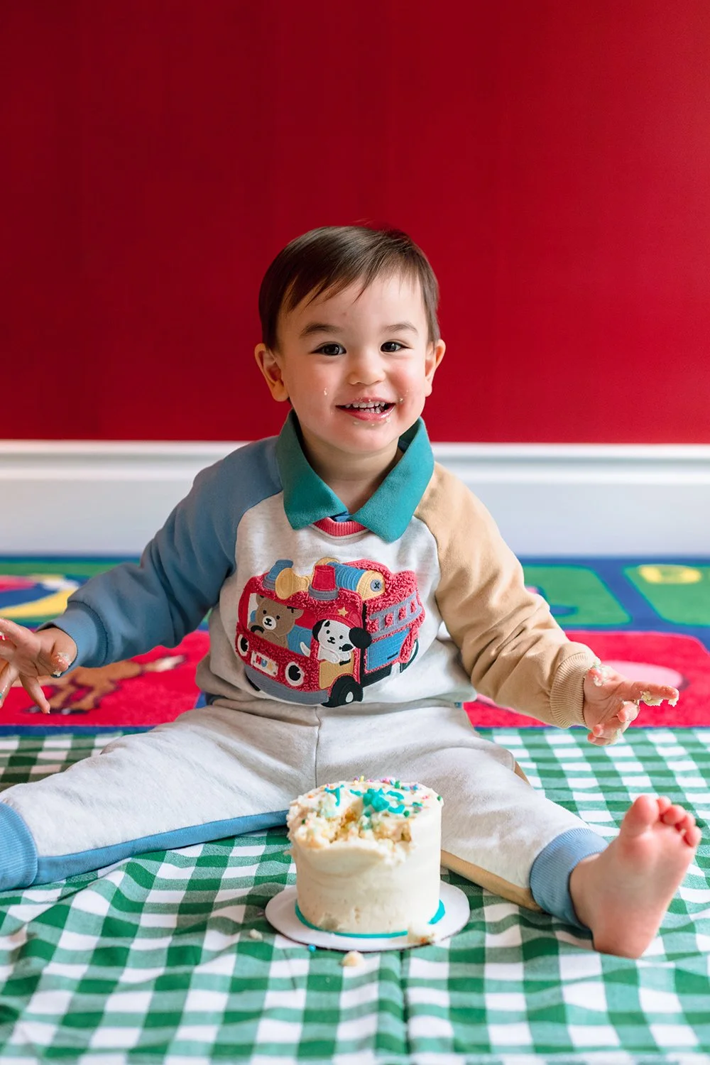 Smiling young boy celebrating birthday with cake, sitting on a checkered blanket in front of a red wall.