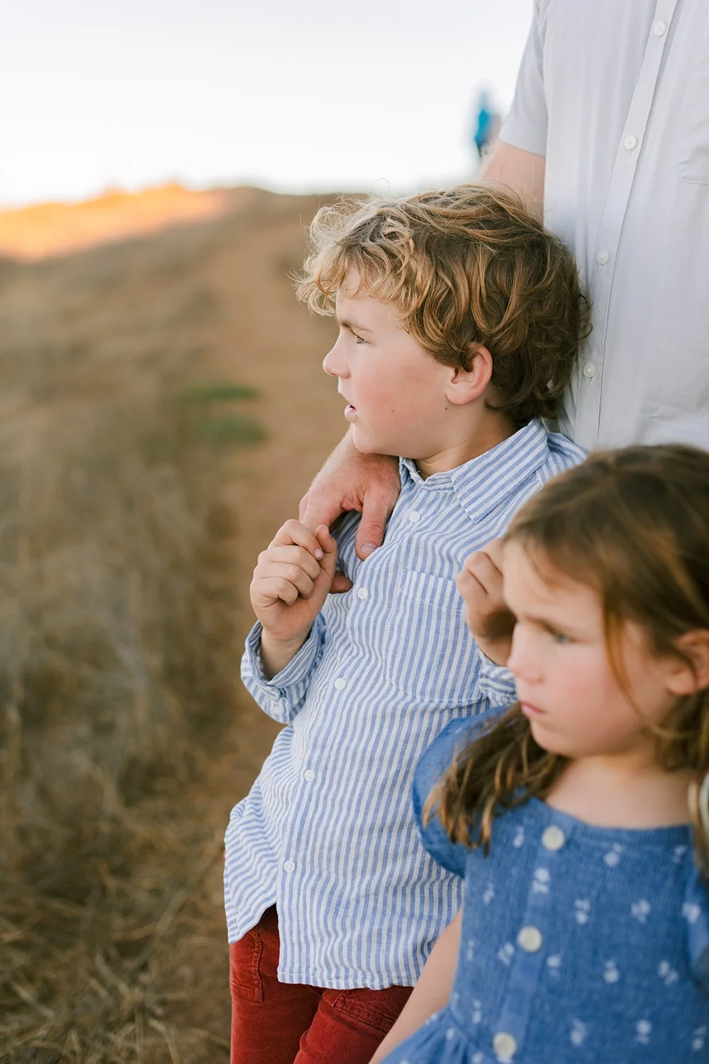A boy with curly light brown hair and a girl with straight brown hair standing outdoors, with an adult's arm around the boy. The background shows a blurred landscape with a sunrise or sunset.