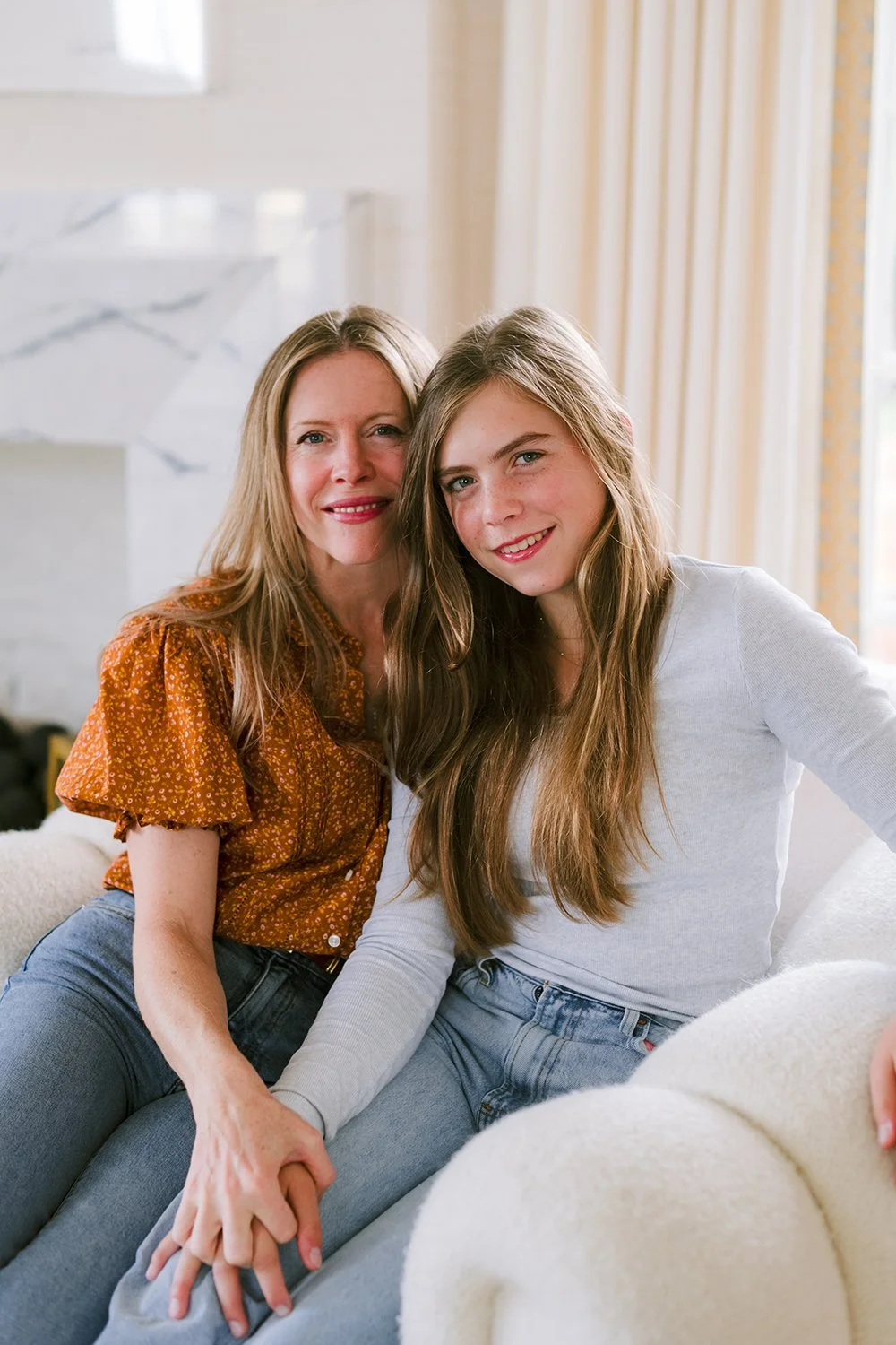 A mother and daughter smiling and sitting close together in a cozy, sunlit living room.