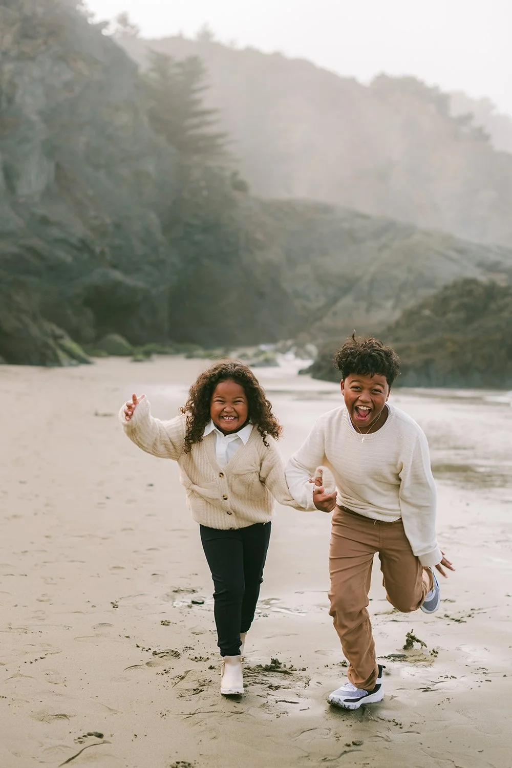 Two children, a girl and a boy, joyfully running and playing on the beach with rocky cliffs and trees in the background.