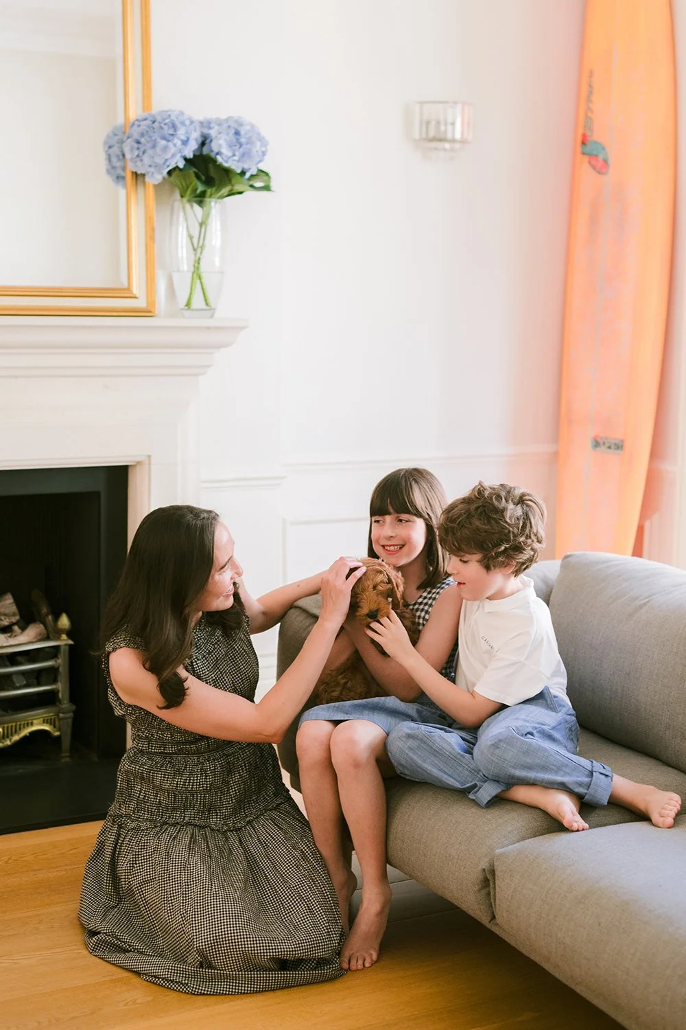 A woman and two children sitting on a couch playing with a brown puppy in a bright living room.