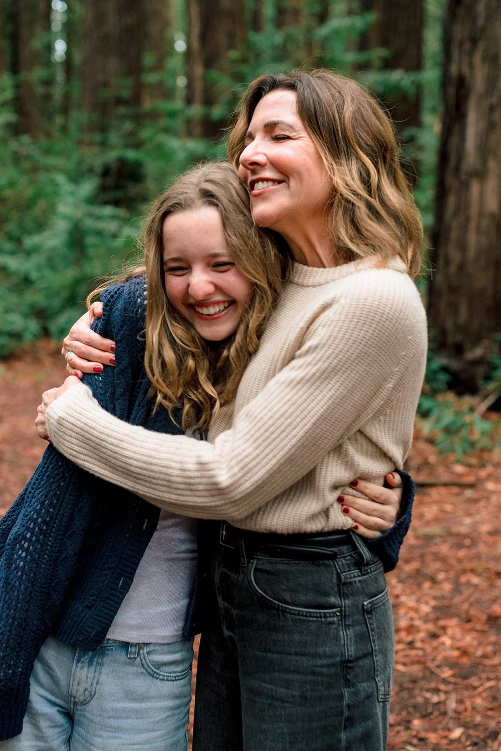 A woman and a girl hugging and smiling in a forest with tall trees and green foliage in the background.