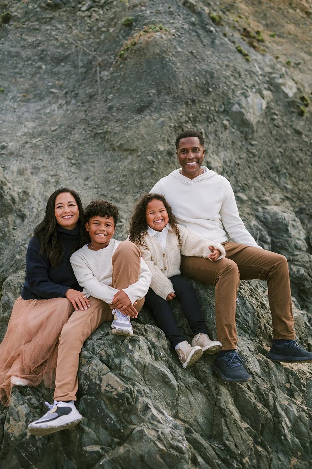 A family of four sitting on rocks outdoors, smiling at the camera, with rocky terrain in the background.