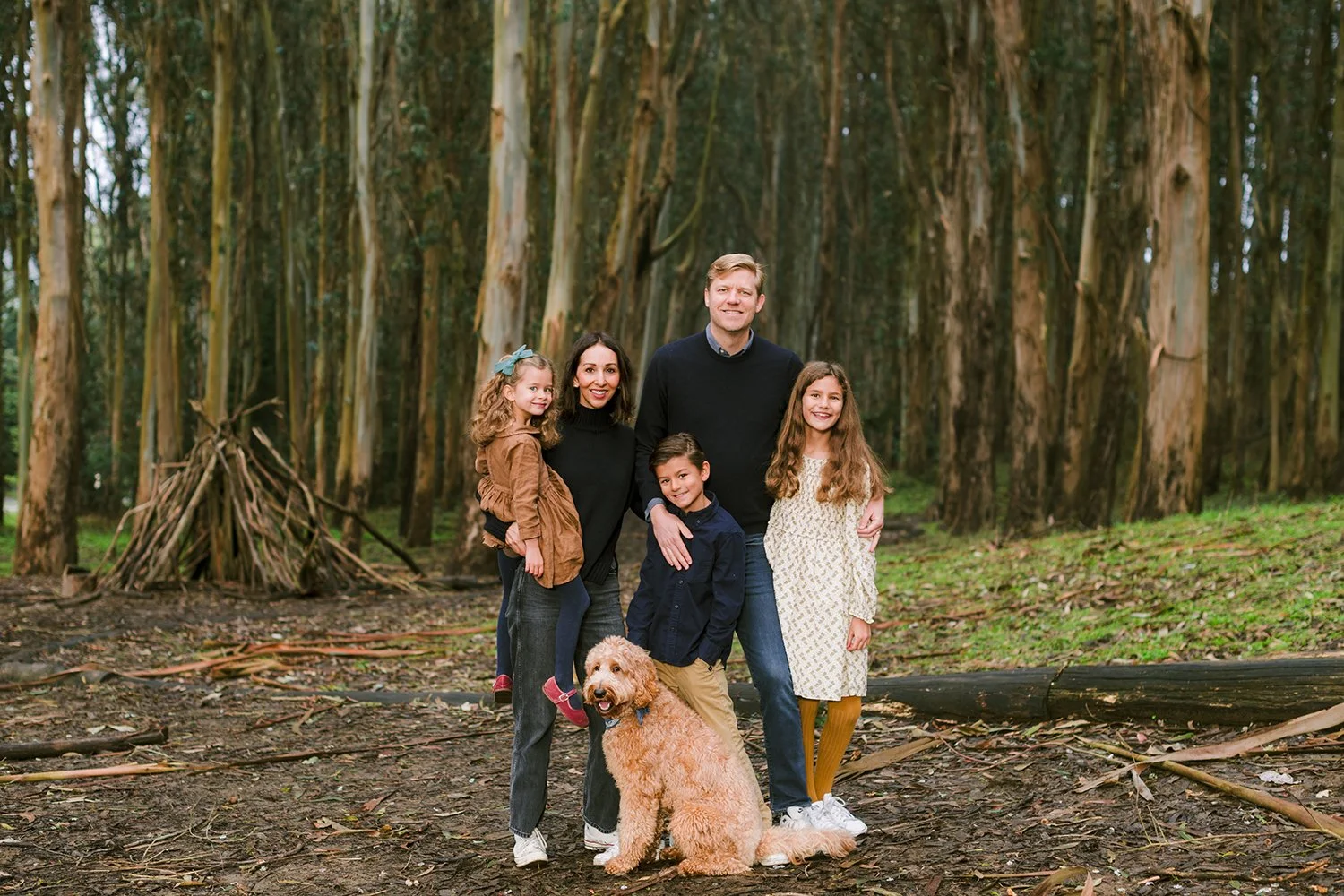 Family of five with a dog standing in a forest with tall trees and green foliage.