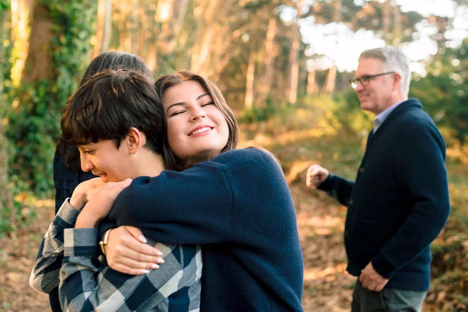 Four people hugging outdoors in a forested area with sunlight filtering through the trees.