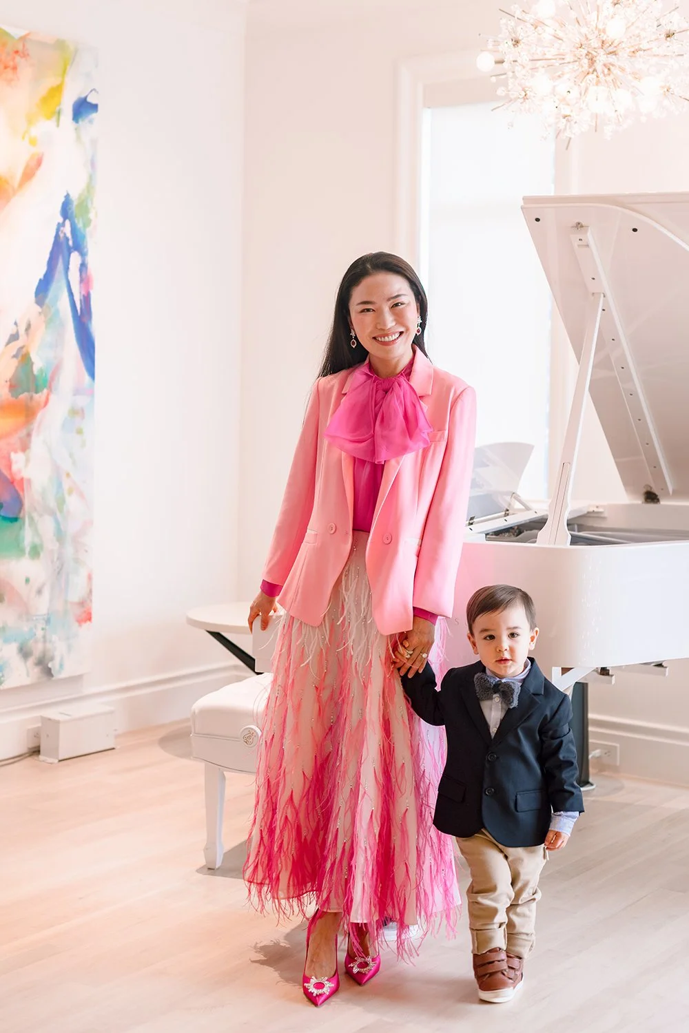 A woman in a pink blazer and skirt smiling and holding hands with a young boy dressed in a navy blazer, beige pants, and brown shoes, standing in a bright room with a white grand piano and a colorful abstract painting on the wall.