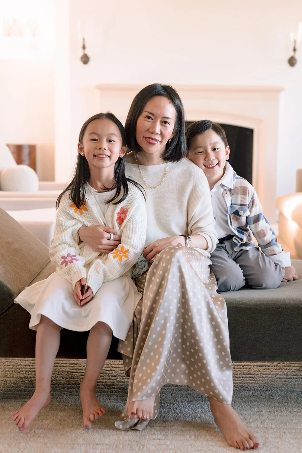 A woman sitting on a couch with a young girl and boy on either side of her, all smiling at the camera, in a cozy living room.
