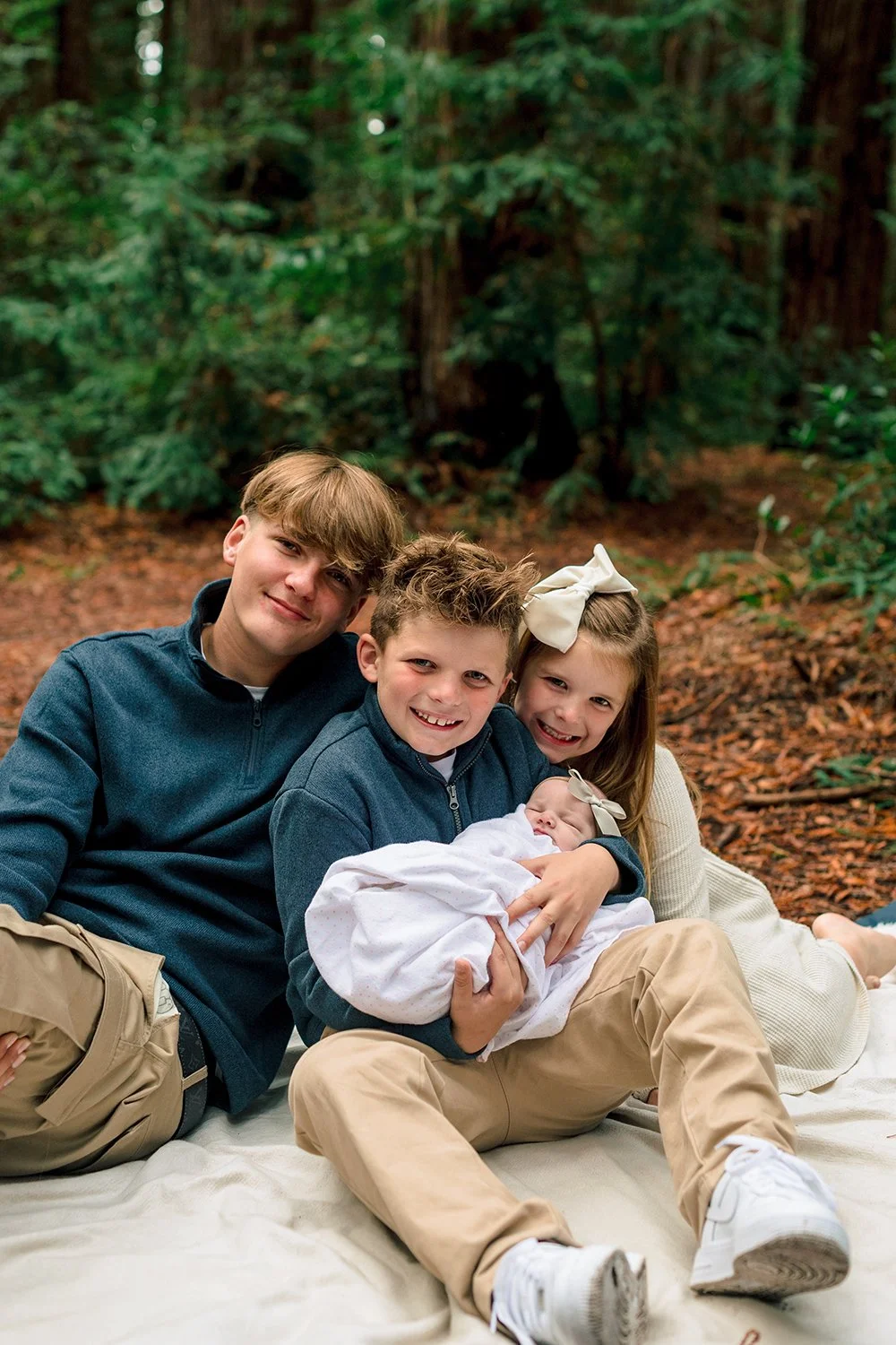 Family of four, including a newborn, sitting on a blanket in a forest