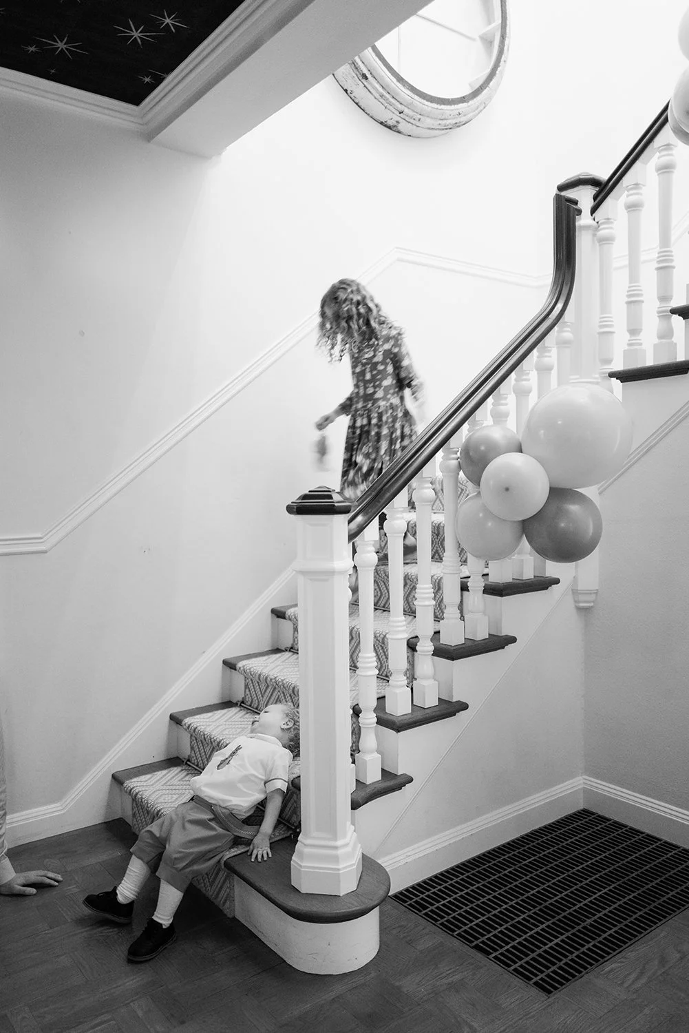 A woman walking down a staircase decorated with balloons, while a child is lying on the bottom step, appearing to be asleep or resting.