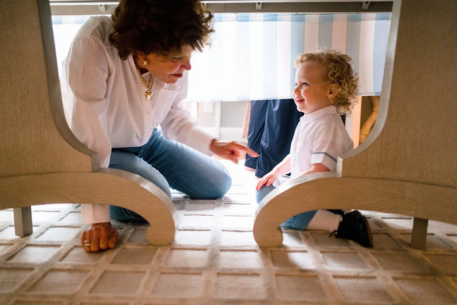 A woman and a young girl sitting on the floor under a bed, engaged in a conversation, as seen from underneath the bed. The woman is smiling and pointing at the girl, who is looking at her and smiling. The scene is well-lit with natural light.