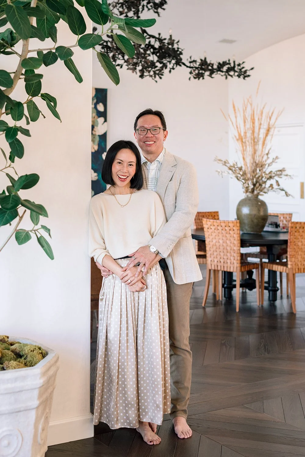 A smiling couple standing barefoot in a bright, modern dining room, with the woman wearing a beige sweater and polka-dot skirt and the man in a light blazer and trousers, amid interior decor including a large vase with dried branches and woven chairs