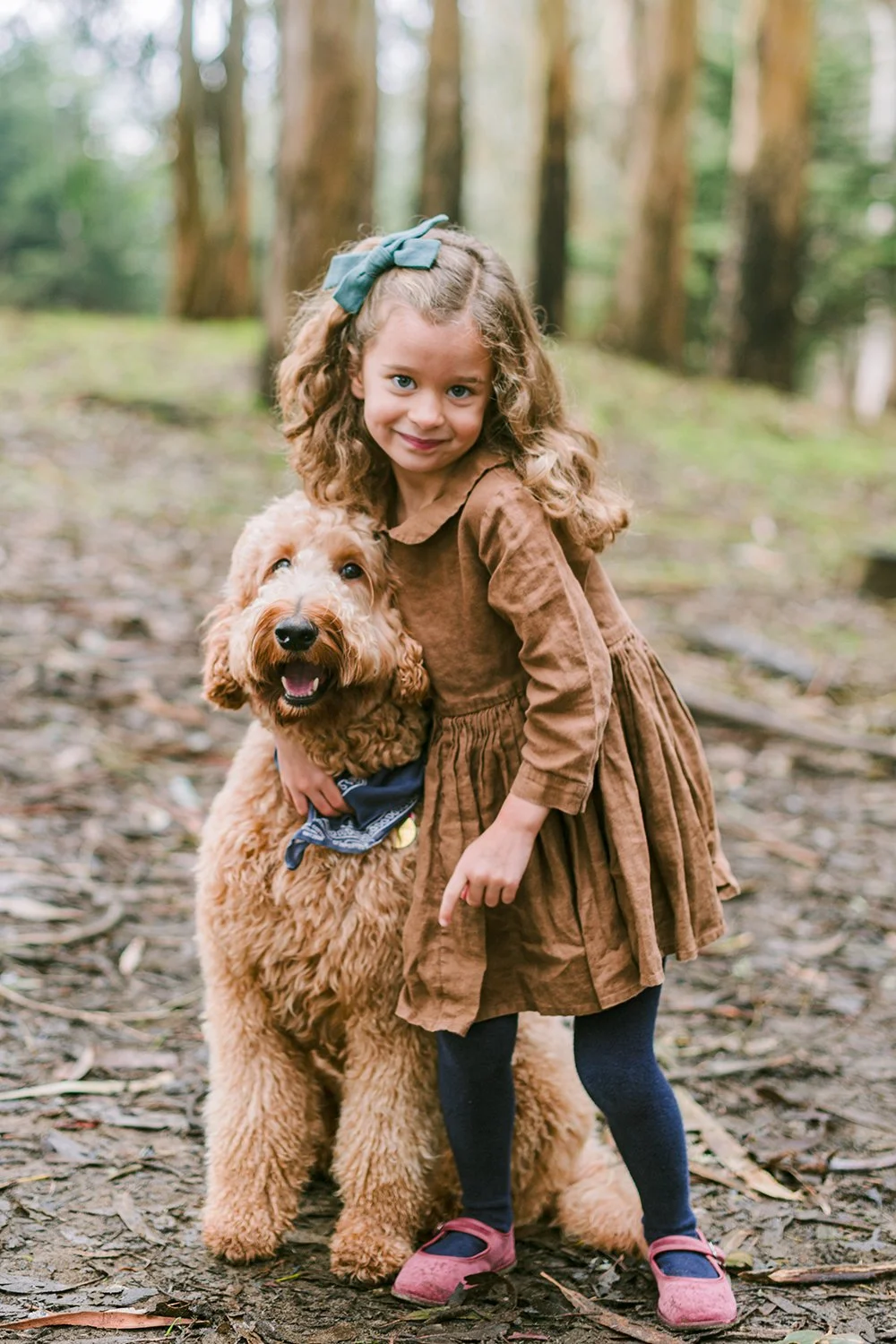 A young girl with curly hair and a blue bow, dressed in a brown dress with blue tights and pink shoes, stands outdoors in a wooded area, hugging a large, fluffy, golden retriever dog with a blue bandana around its neck.