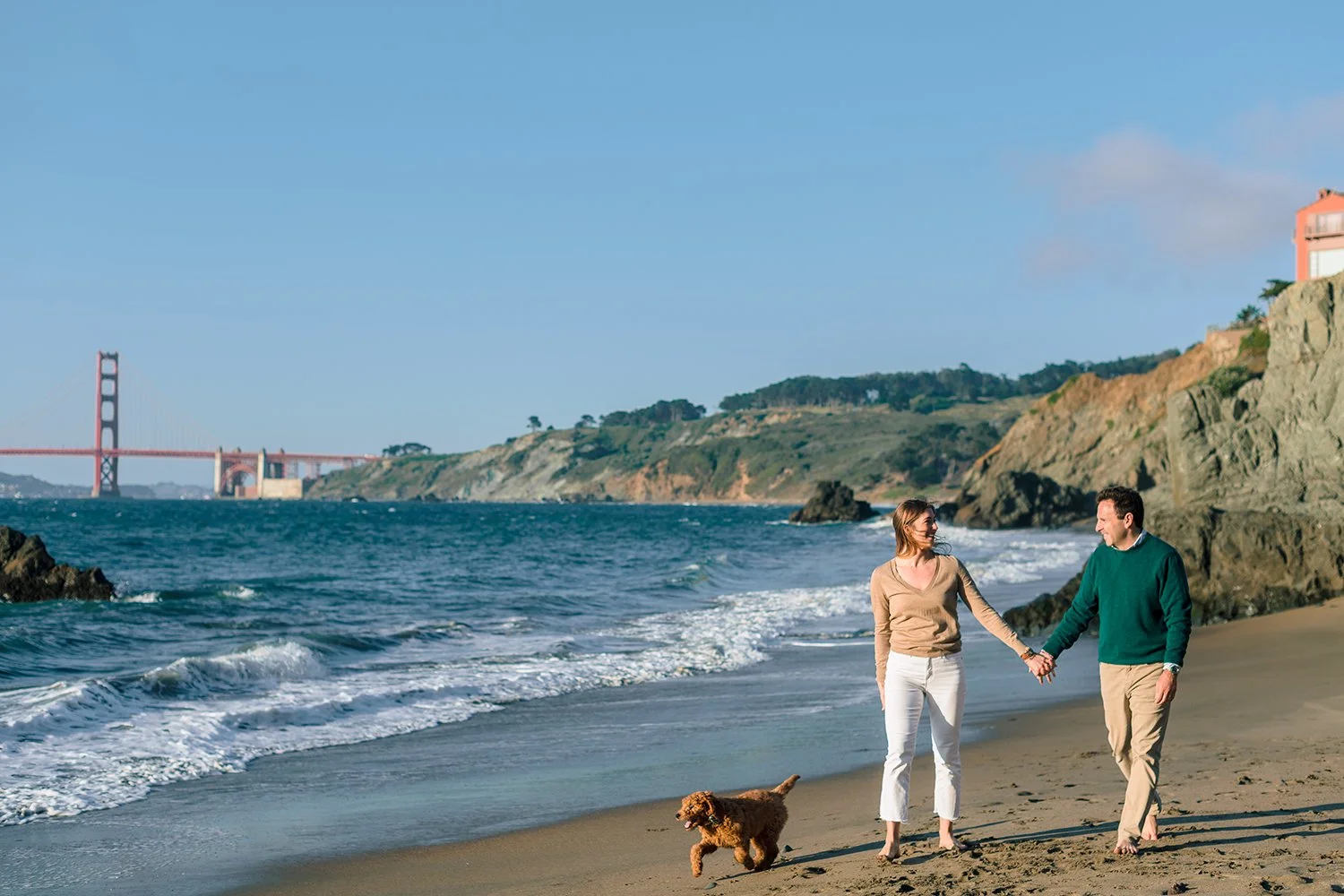 A couple walking hand-in-hand with their dog on a sandy beach near rocky cliffs, with the Golden Gate Bridge in the background on a clear day.