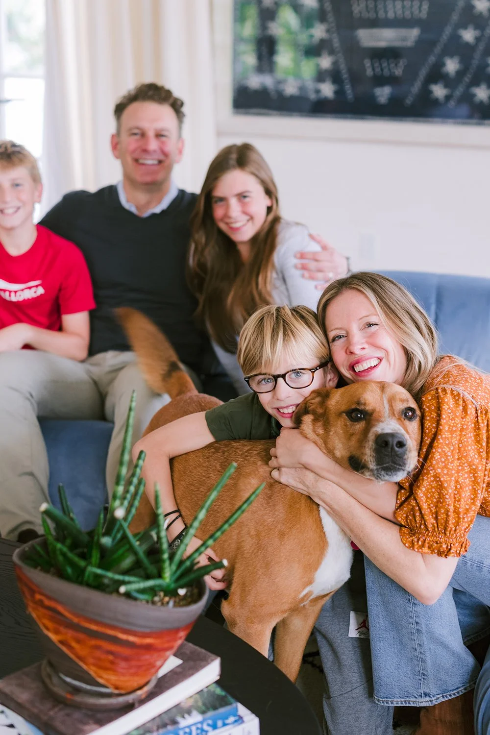 Family gathered in living room, smiling and hugging their dog, with a potted plant and books on a table in the foreground.