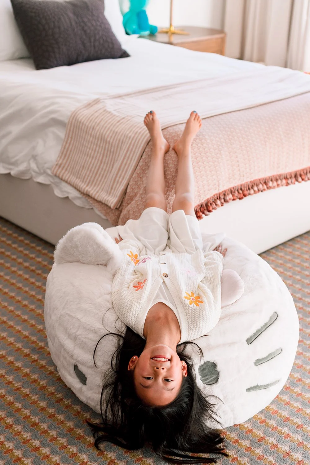 A smiling young girl lying upside down on a plush, animal-shaped pillow in a cozy bedroom.
