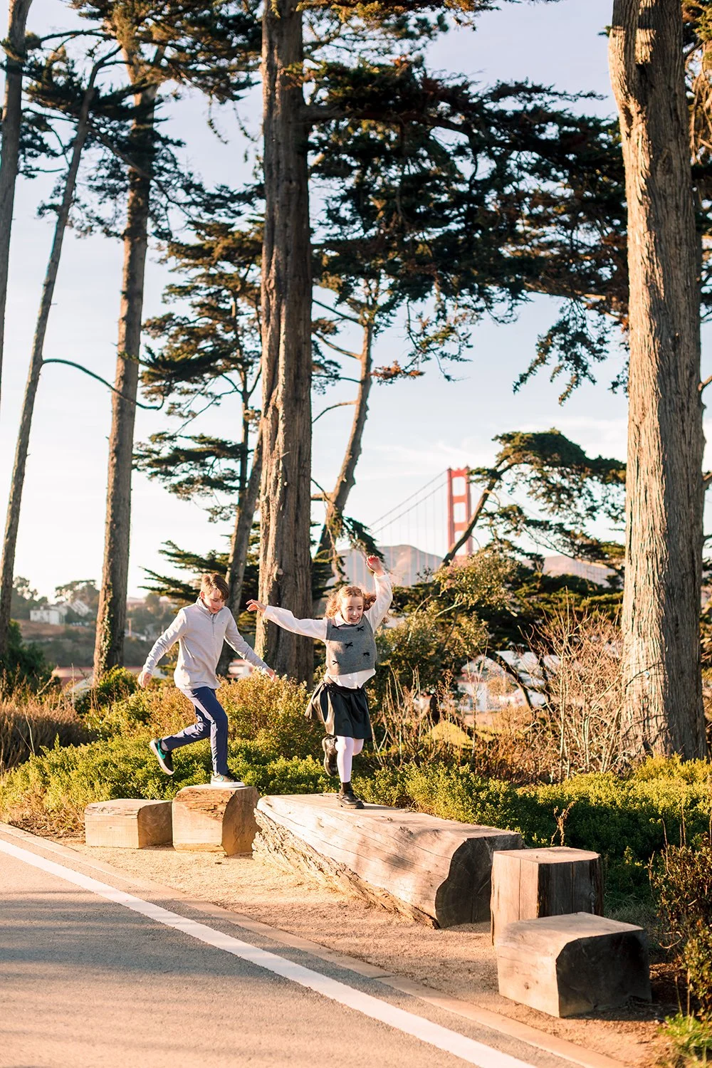 Two children, a boy and a girl, are playing on large wooden logs used as outdoor stepping stones next to a paved path. The girl is jumping, and the boy is stepping onto the logs. Tall trees and the Golden Gate Bridge are visible in the background dur
