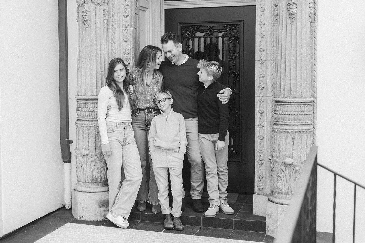 A black and white photo of a family standing together in front of an ornate door; three children and two adults sharing a happy moment, smiling and hugging.
