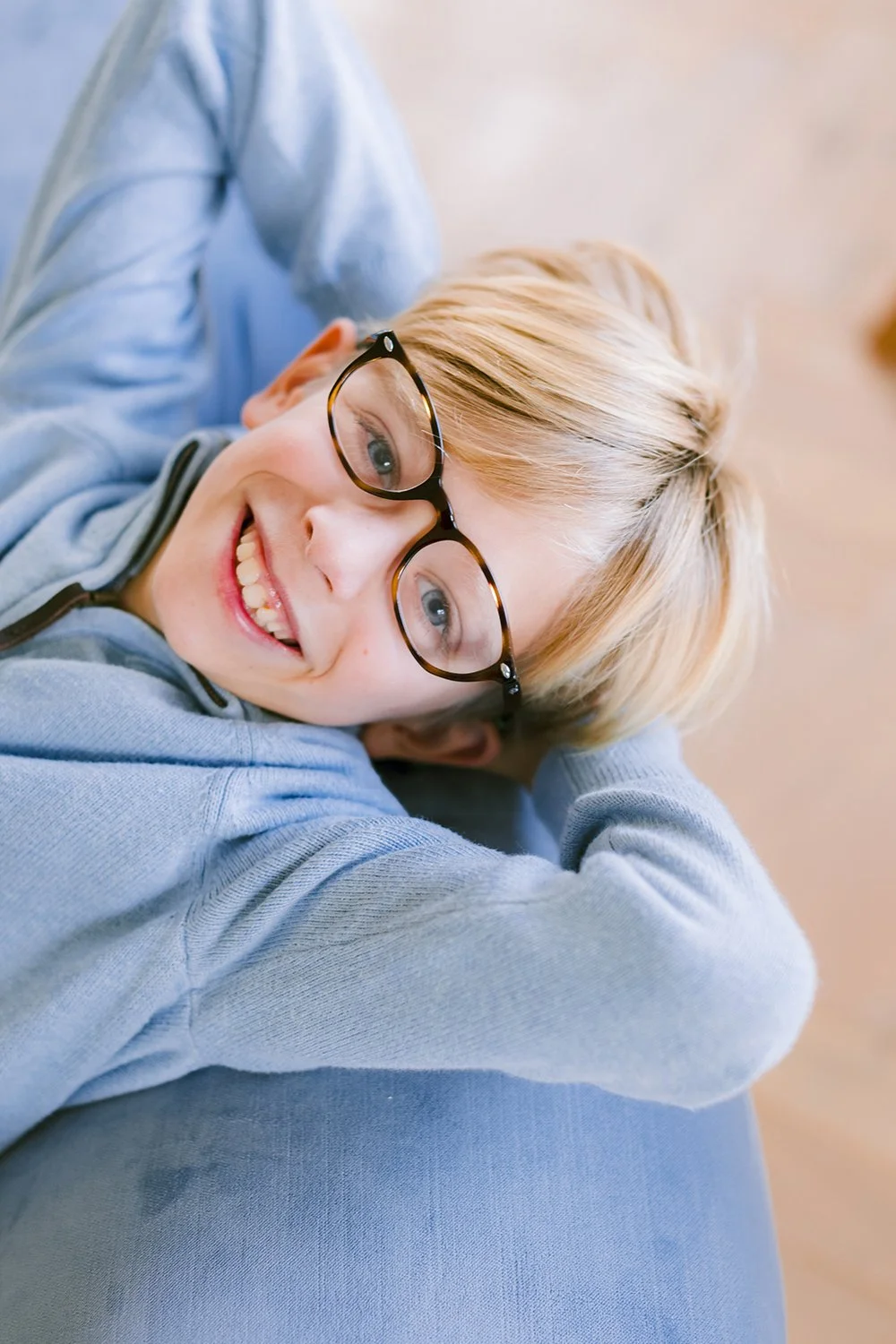 Close-up of a smiling boy with blonde hair and glasses, lying on a couch with a light blue hoodie.