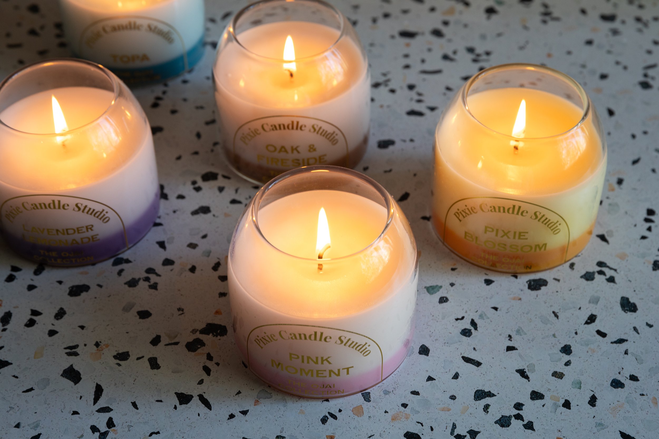 Several white candles in glass holders burning on a speckled tabletop. The candles are labeled with white and gold stickers, indicating scents such as lavender lemonade, pink moment, and oak & fireside.