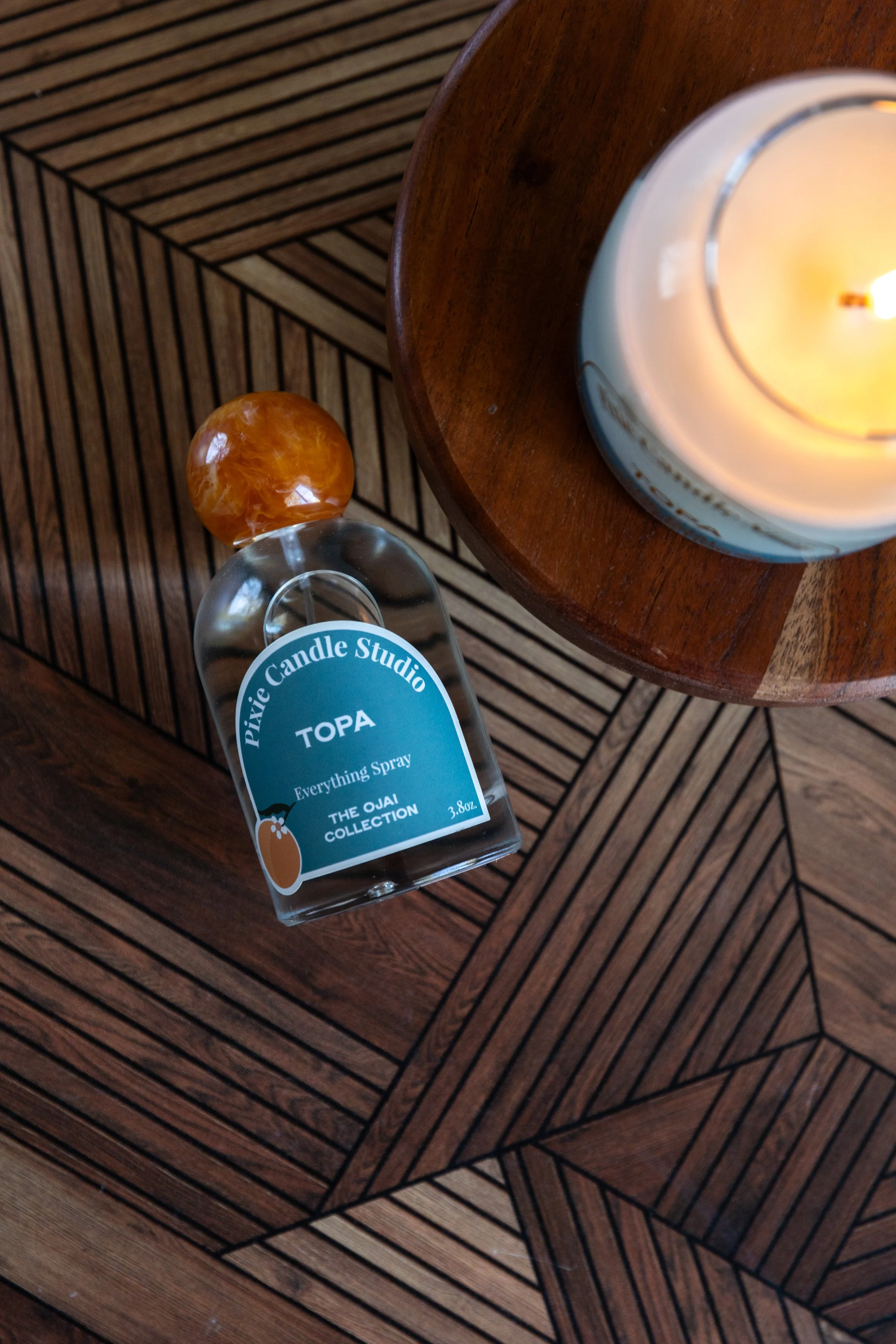 A lit white candle in a jar placed on a wooden table, next to a glass bottle with a brown and yellow marbled ball on top, and a wooden box with a geometric pattern.